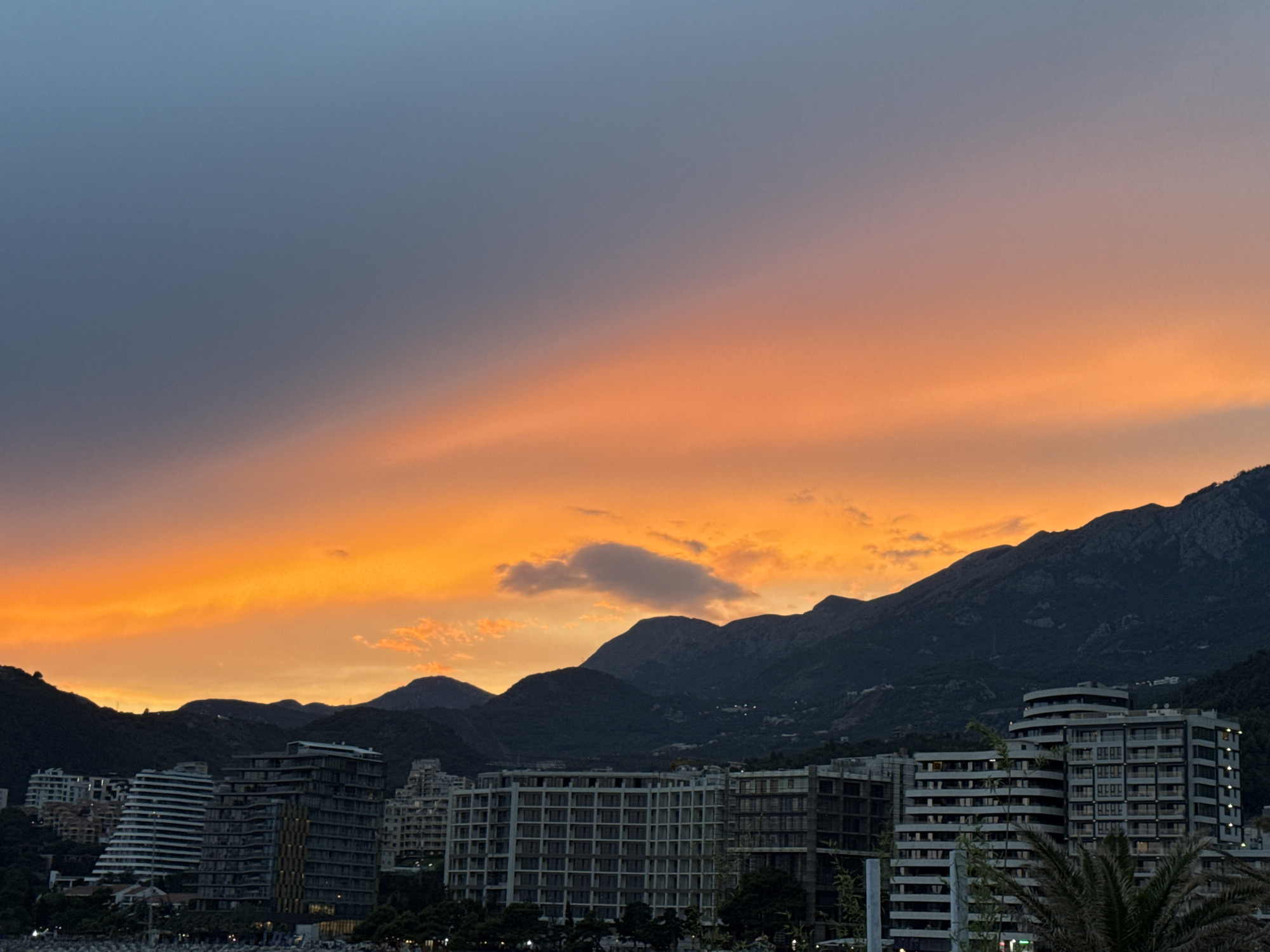 The image depicts a coastal city scene at dusk, likely Budva, Montenegro, with a dramatic sunset painting the sky. The foreground features several modern, multi-story buildings, some with balconies and extensive window systems, appearing to be hotels or residential complexes. The architecture is contemporary, with clean lines and varying facade designs. Some of these buildings are illuminated from within, suggesting occupancy.
Behind the buildings, a range of dark, imposing mountains rise, their slopes covered in lush vegetation, which is silhouetted against the vibrant sky. The sky itself is a spectacle of color, transitioning from a deep, dusky blue at the top to a brilliant, fiery orange and yellow near the horizon, indicative of the setting sun. Wispy clouds catch the light, adding texture and depth to the atmospheric display. One larger, isolated cloud is visible in the mid-sky.
The lower portion of the image shows hints of greenery, possibly trees or palm fronds, and what appear to be outdoor seating areas or walkways along the waterfront, though these are partially obscured. The overall impression is one of a developed coastal area harmonizing with a natural mountainous backdrop, illuminated by the striking beauty of a sunset. There are no visible people or ongoing actions, presenting a still, scenic view of the city at the end of the day.