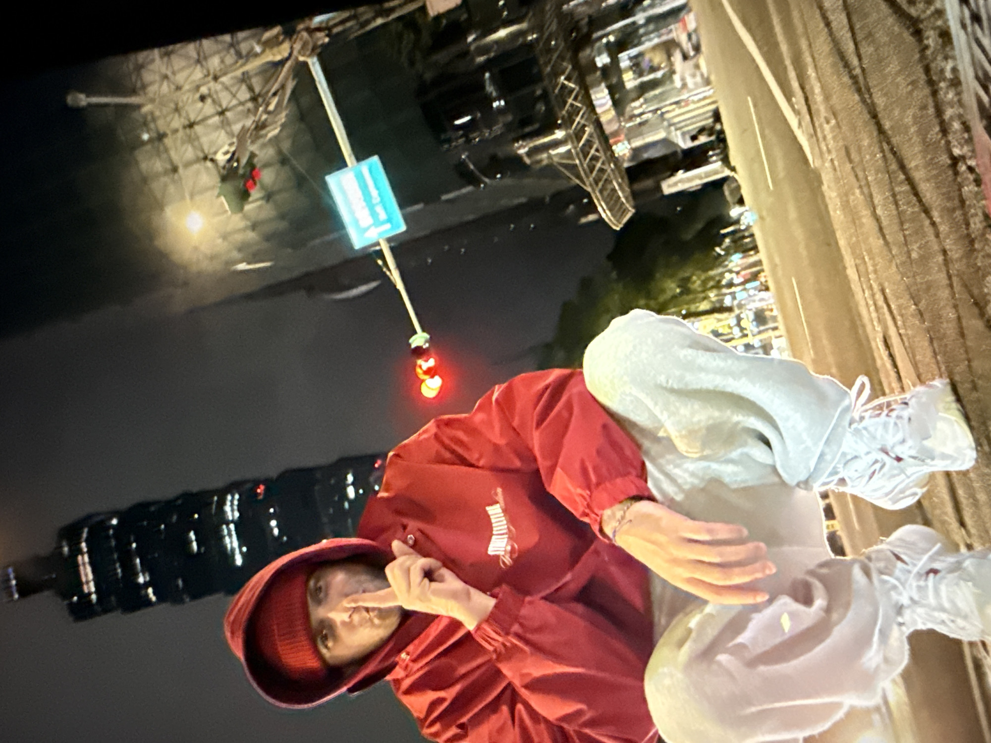 A low-angle, night-time, outdoor medium shot shows a person squatting on a street in Taipei, Taiwan. The person, seen from the waist down and including their head, is wearing a red beanie, a red hooded jacket with the text "STREET CULTURE" in white embroidered on the chest, light grey sweatpants, and white sneakers. The person has their index finger to their lips, signaling silence. Behind the person, a tall skyscraper, identifiable as Taipei 101, is visible in the background, illuminated by night lights. Traffic lights, street signs, and the blurry lights of cars and city buildings are also present, indicating an urban environment. The street surface is wet, possibly from rain, and reflects the surrounding lights.