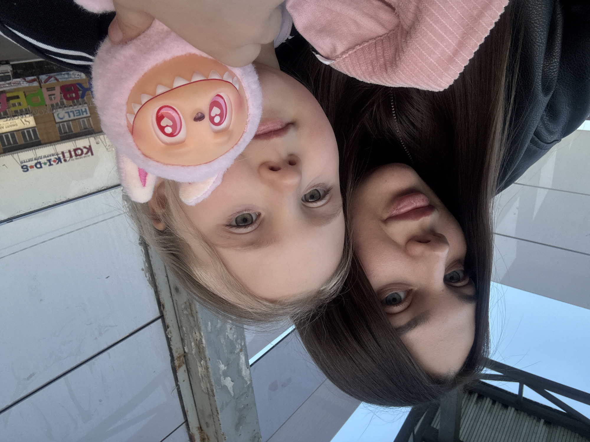 The image captures a selfie taken in the city of Chaikovsky, Russia. The focus is on a fair-skinned woman with long, dark brown hair and a young girl with blonde hair. The woman is wearing a black leather jacket and the girl is wearing a pink hoodie. The girl is holding a pink plush toy with heart-shaped eyes and a toothy grin. The background includes a building with signs in Russian, including what appears to be a children's center named "Karik-Kids". The overall setting suggests an urban environment.