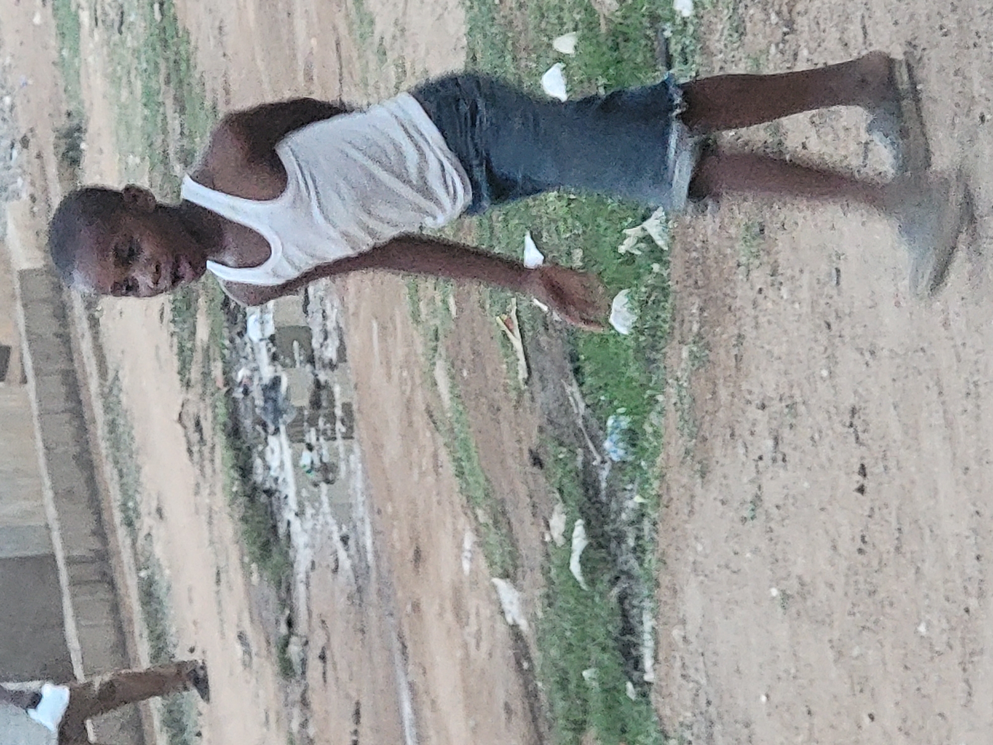 The image depicts a young person standing on a dirt path in Sabon Tasha, Nigeria. The individual is wearing a white tank top, dark-colored shorts, and light-colored shoes. The surroundings include patches of green vegetation, scattered pieces of white debris, and, in the background, a cluster of buildings, indicating a populated or residential area. The scene conveys an outdoor environment in an urban or semi-urban setting.