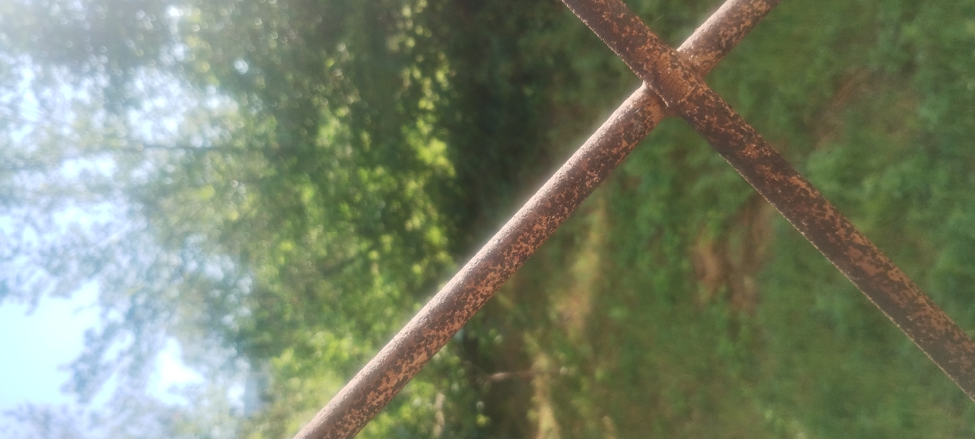 The image shows a close-up of two rusty metal bars that form an 'X' shape in the foreground. The bars are textured with patches of brown and orange, indicative of rust. Behind the bars, the background is out of focus, featuring lush green foliage and a bright sky. The bokeh effect suggests a shallow depth of field. The overall scene depicts an outdoor environment, possibly a park or a backyard, on a sunny day. There are no people or discernible structures visible in the image. The image is taken in Zaporizhzhia, Ukraine.