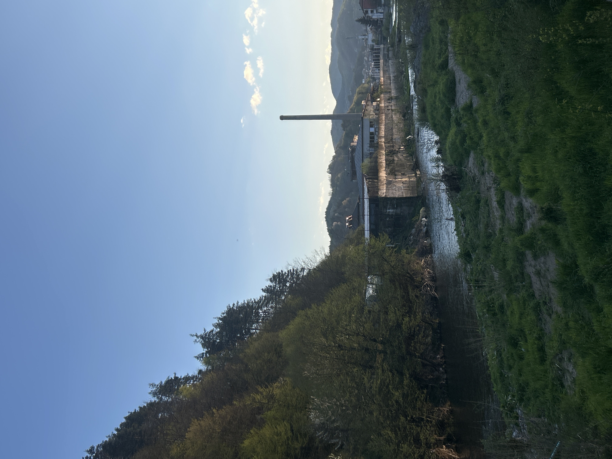This landscape image captures a valley scene, likely in the late afternoon or early morning, under a bright, clear blue sky with a few scattered white clouds. In the mid-ground, a prominent, tall, slender industrial chimney rises into the sky. Adjacent to it stands a large, somewhat dilapidated industrial building or factory with visible windows, suggesting an older structure.
To the right of the factory, a river or canal flows towards the foreground, its banks covered in lush green grass and varying shades of green foliage. Dense forests and trees line the hillsides, particularly on the left side of the image, with some trees showing lighter green new growth.
In the background, behind the industrial structures, a range of mountains or hills extends across the horizon, with hints of a small town or village nestled in the valley. The sunlight creates a soft glow and subtle haze over the distant landscape and parts of the factory. No people are visible, and there is no apparent activity taking place. The scene offers a view of a blend of natural environment with industrial elements, typical of a town situated in a mountainous river valley like Zlatograd, Bulgaria. No text is visible in the image.