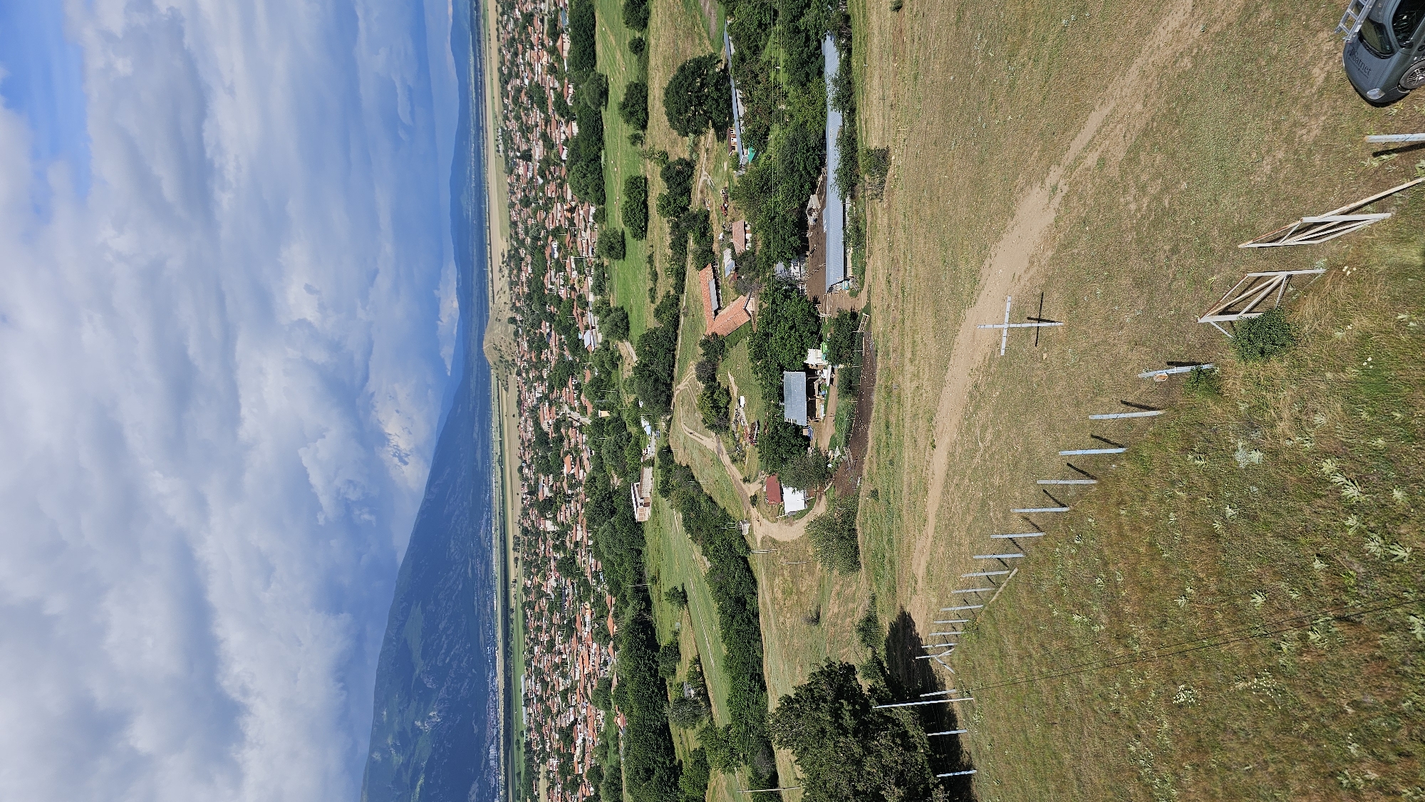 The image is a high-angle aerial view of a rural landscape in or near Dabene, Bulgaria. In the foreground, a dry, grassy hillside is marked by a metal fence and a dirt path. A wooden cross stands near the path. Further down the slope, a small cluster of farm buildings with red tile roofs are visible, surrounded by trees. Beyond the farm buildings, a dense village with numerous houses, also with red tile roofs, stretches across the middle ground. The village is nestled within a verdant, green landscape dotted with trees. In the background, a range of mountains forms a dramatic backdrop, partially obscured by a cloudy sky. The overall impression is of a peaceful, agrarian community in a mountainous region under a dynamic sky.