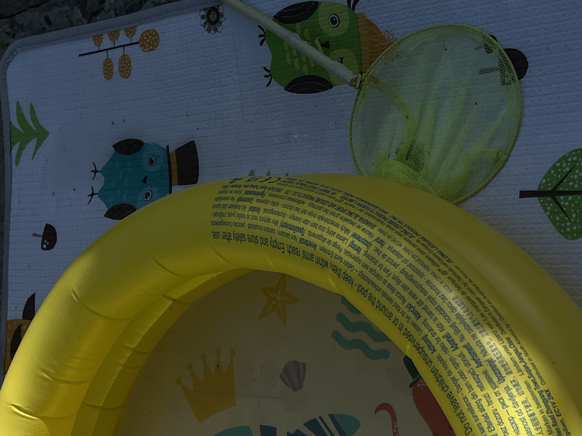 The image shows an inflated yellow kiddie pool placed on a blue and white mat with owl and floral illustrations. A green-netted fishing scoop with a white handle rests on the mat, partially overlapping the pool. Text in multiple languages is visible on the yellow pool. The setting appears to be outdoors, possibly a backyard or patio, as indicated by the textured ground visible in the background. There are no people or animals visible in the image. The location is not specified as Novopokrovskaya, Russia; the text on the pool does not provide location information for the image itself.