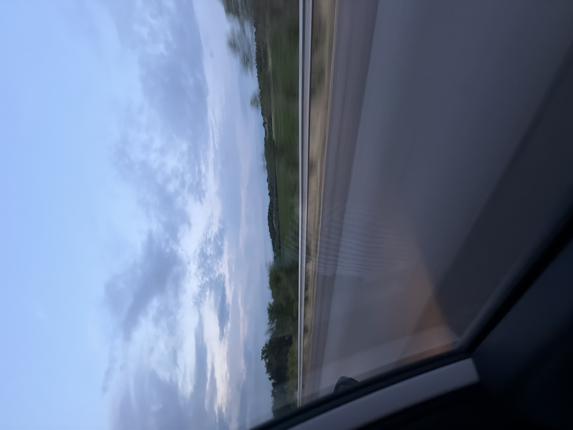 The image presents a sideways view from inside a moving vehicle, looking out through a side window at a landscape under a cloudy sky. The sky, occupying the upper left portion of the frame, is a blend of light blue and soft grey clouds, suggesting either an overcast day, dawn, or dusk. Below the sky, a verdant landscape with green fields and a distant treeline appears blurred horizontally, indicating the vehicle's motion. A grey guardrail or barrier runs across the mid-ground, separating the landscape from what seems to be a road or shoulder, also exhibiting motion blur. The lower right section of the image reveals the dark frame of the car window and a glimpse of the vehicle's interior, with faint reflections visible on the glass. There are no people or discernible text in the image. The scene captures an event of travel through a rural area, with the provided context indicating a location near Kalugerovo, Bulgaria.