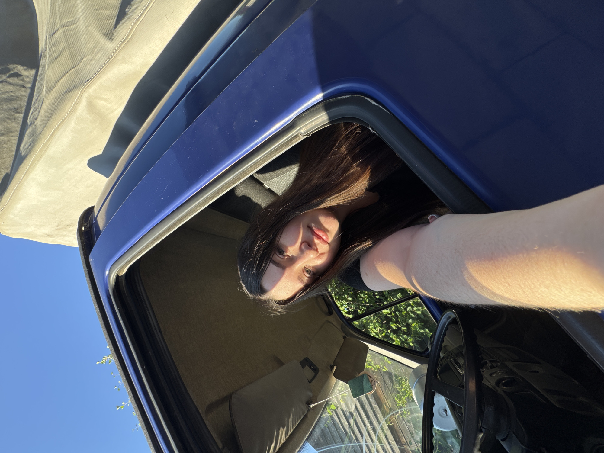 A young woman with long, dark hair is looking out of the passenger side window of a blue vehicle. She is wearing a black shirt. Her right arm is extended out of the window, casting a shadow on the exterior of the vehicle. The interior of the vehicle is visible, including the steering wheel and dashboard. Outside the vehicle, lush green foliage and a clear blue sky are visible. The vehicle appears to be parked outdoors, possibly in a residential area, given the presence of a fence-like structure in the background. The lighting suggests it is late afternoon or early evening, with the sun casting long shadows. The image was taken in the city of Krasnokamsk, Russia.
