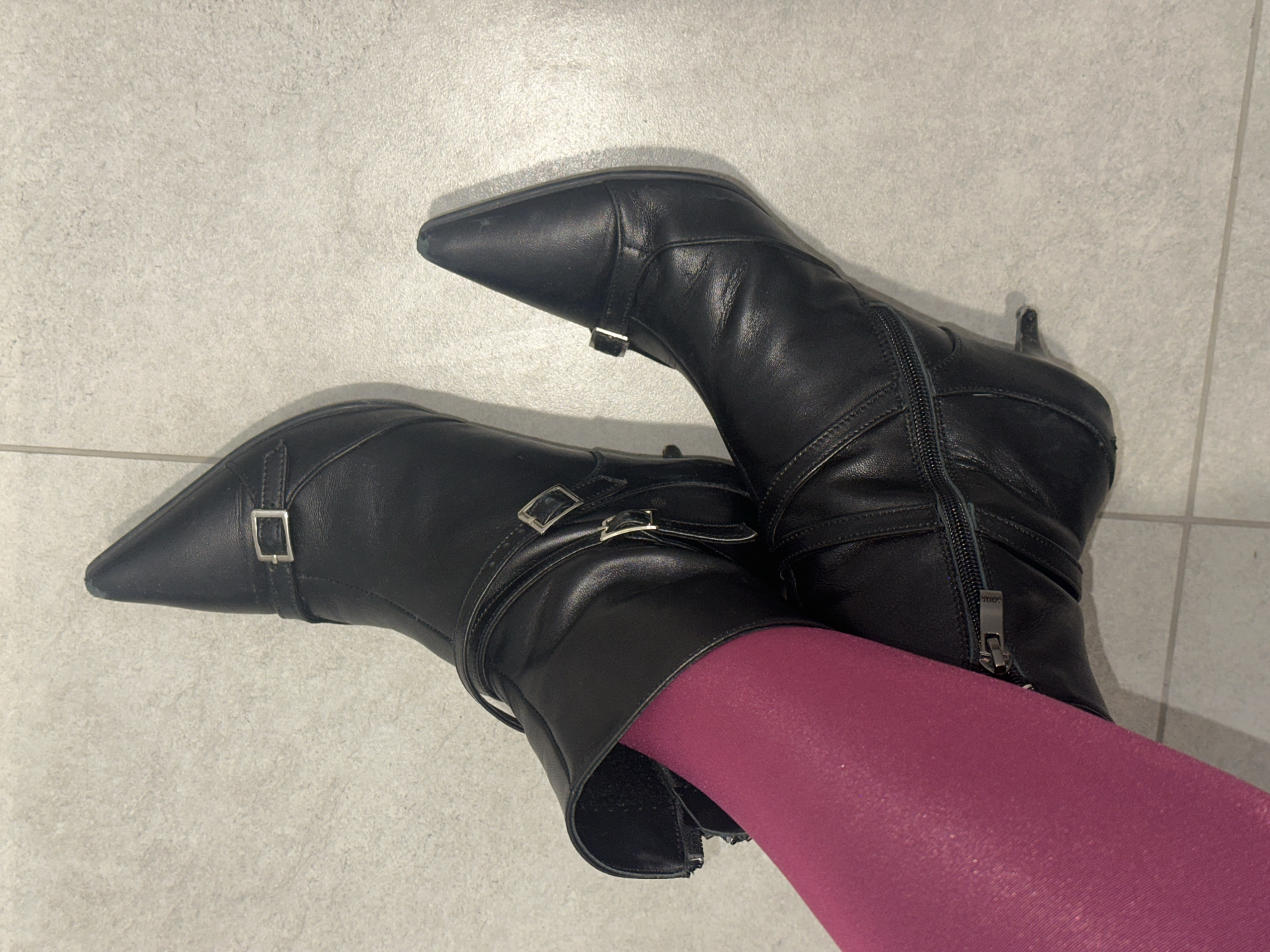 Two black leather pointed-toe ankle boots with buckle details are displayed on a light gray tiled floor. The boots are worn by a person wearing bright magenta tights. The left boot is positioned in the foreground and is partially unzipped, revealing the magenta tights. The right boot is behind the left, with its toe pointing upwards. The background is a neutral gray tiled surface.
