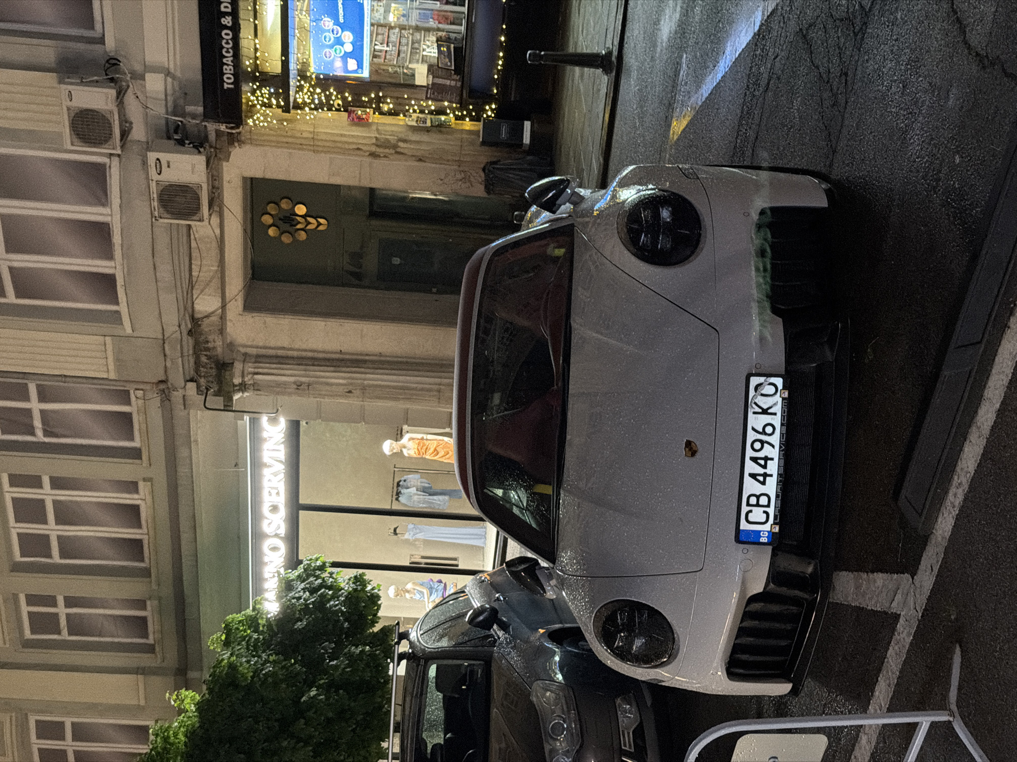 The image captures a nighttime street scene in a city, likely Sofia, Bulgaria. A light grey Porsche sports car is prominently parked on the wet street, suggesting recent rain. Its front fascia is visible, showing the headlights and details of the bumper, all covered in small water droplets. The vehicle's license plate, located on the front bumper, clearly displays "CB 4496 KO" and includes a "BG" country code sticker, confirming its Bulgarian registration. Below the main registration, faint, small text appears to be a website address: "IASURAITSERVICE.COM".
To the left of the Porsche, a portion of another dark-colored vehicle is visible. The street and curb are dark and reflective from the wet conditions, with subtle road markings visible. A white metal barrier or rack partially extends into the foreground on the right.
Along the left side of the street, multiple buildings rise, with one featuring several distinct windows. Two outdoor air conditioning units are mounted on its facade. Further along, a brightly illuminated shopfront draws attention, featuring a vertical glowing sign that reads "VALENO SERVIC". Inside this shop's large window, several mannequins, dressed in various garments, are on display. Above this establishment, another horizontal shop sign is partially visible, reading "TOBACCO & D...". The area is adorned with strings of small, warm yellow decorative lights. A green tree or bush is situated near the base of the buildings. The entire scene is lit by a combination of streetlights and the glow emanating from the shop windows.