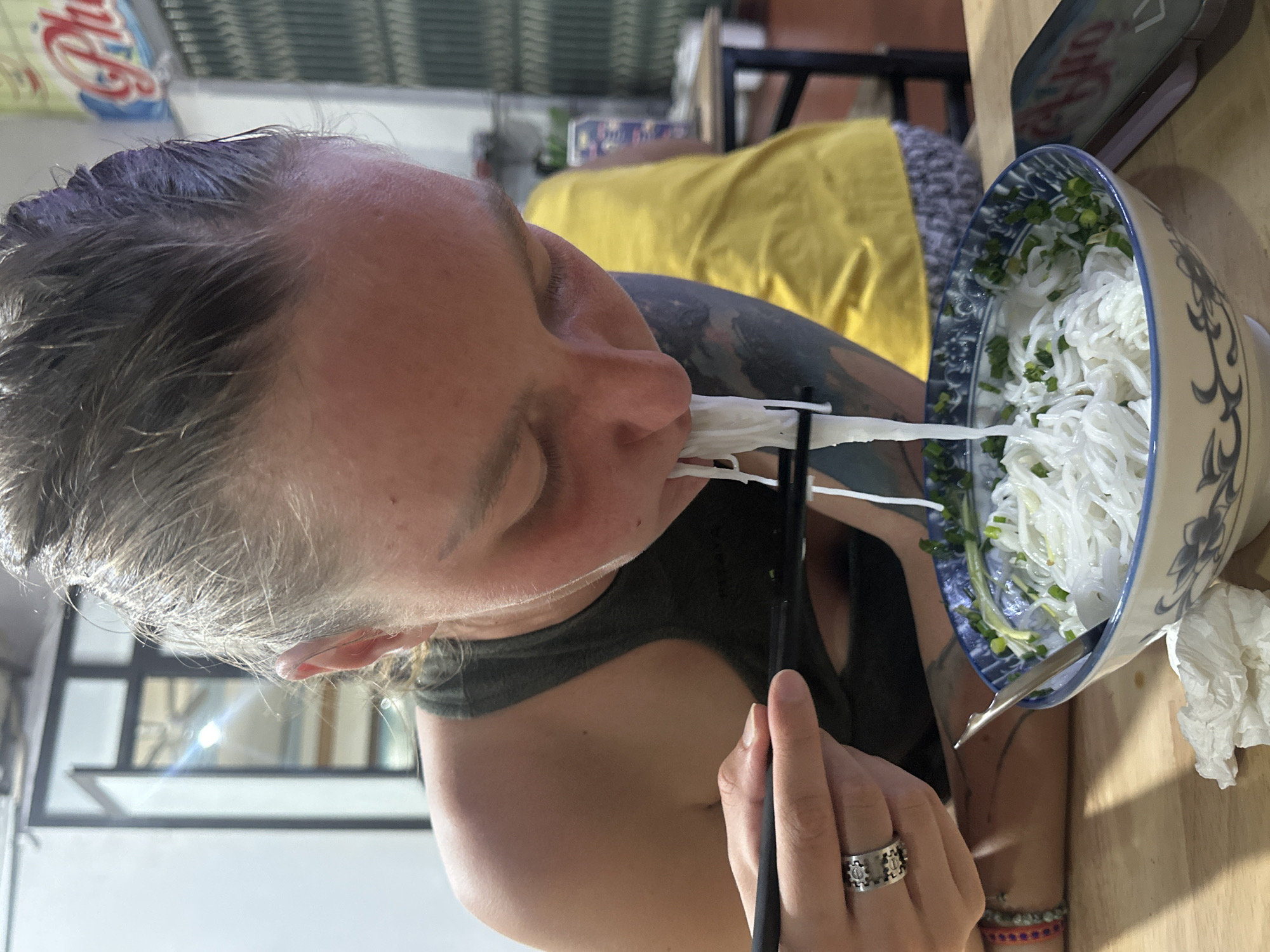 A light-skinned individual with hair pulled back is depicted in a medium shot, actively consuming white vermicelli noodles from a patterned ceramic bowl using black chopsticks held in their right hand. The individual wears a dark green tank top and displays a dark tattoo on the upper left arm. A silver-toned ring with a mechanical design is visible on the right ring finger, and a multi-colored beaded bracelet is on the left wrist. The white noodles are partially submerged in a broth and topped with chopped green onions. A metal spoon rests within the bowl. Adjacent to the bowl on the light wooden table surface are a crumpled white paper napkin and a light-colored smartphone with a screen reflecting red text. In the out-of-focus background, a second person is seated, wearing a yellow shirt and patterned lower garment. Architectural features include a white wall, a dark-framed window or door reflecting a bright light source, and a sign with red and blue Vietnamese text, including the partial word "Phở." Additional background elements consist of vertical slatted structures and simple tables and chairs, indicating an indoor eatery in Trung Tam Ward, Vietnam.