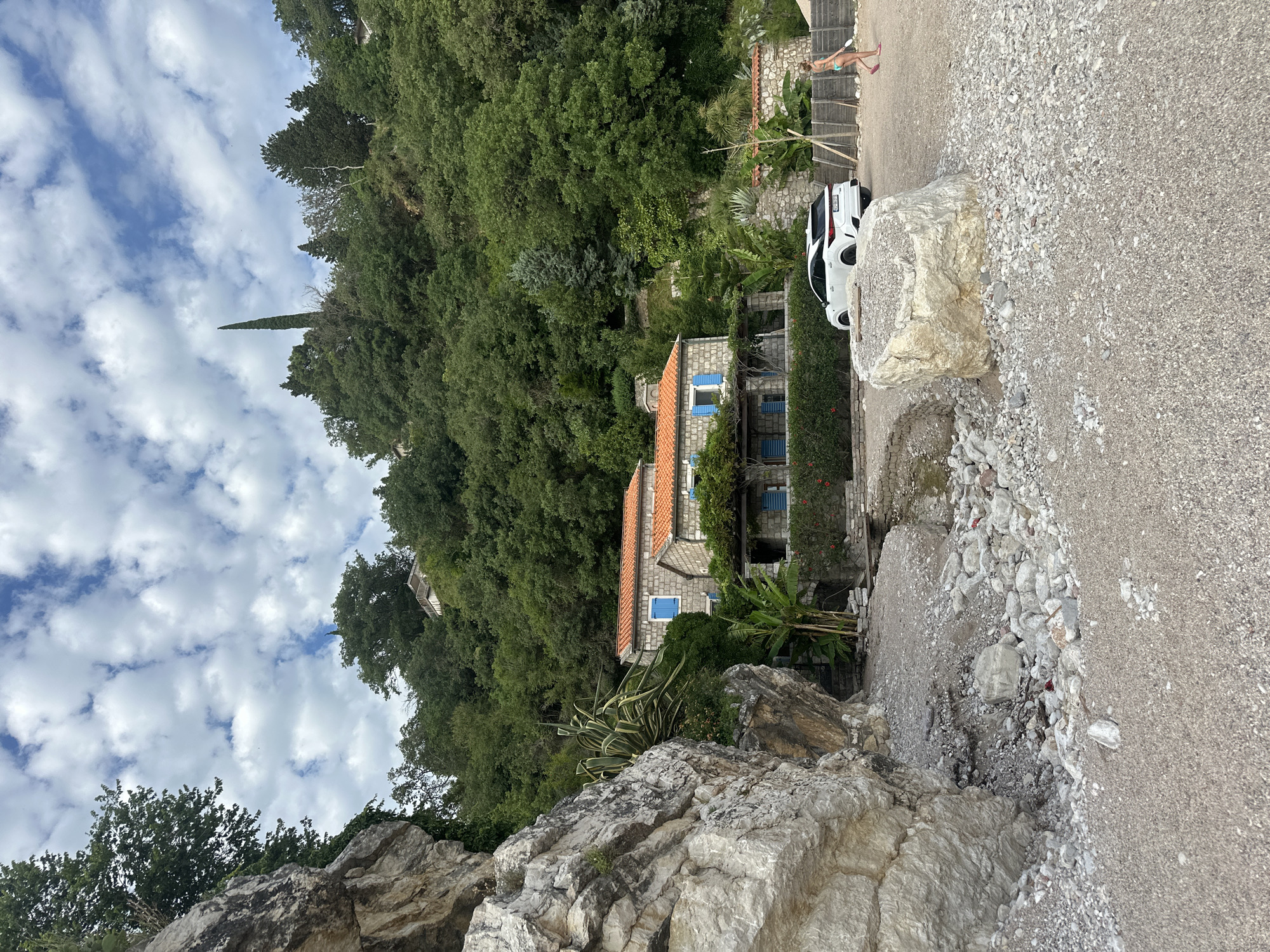 This image depicts a coastal scene in Mirište, Montenegro, characterized by a gravel beach, structures, and a densely vegetated hillside under a partially cloudy sky.
In the foreground, a light brown and grey gravel beach occupies the lower half of the frame. On the left, a large, light-colored rocky cliff face rises vertically, partially obscuring the view behind it. At the base of this cliff, a small, rocky stream bed or drainage channel is visible, containing various grey and white stones. Closer to the center-right foreground, a prominent, light grey-white, irregularly shaped boulder rests on the gravel.
In the midground, towards the right, a white SUV, possibly a Lexus, is parked facing away from the viewer. Its rear license plate, "KT-086," is partially visible. To the right of the parked vehicle, a female individual, wearing a light blue bikini and pink flip-flops, walks from right to left across the gravel. She holds a white object in her right hand. Behind the car, a low stone wall with orange tiled coping separates the gravel area from a manicured hedge and planted vegetation. To the left of the parked car, two stone buildings with orange terracotta roofs and blue window shutters are nestled into the base of the hill. The buildings feature stone facades, and the lower structure has a covered patio area with vines growing over a pergola. Various tropical and Mediterranean plants, including large agave-like succulents and banana plants, grow around these buildings.
The background is dominated by a steep, heavily wooded hill that ascends towards the sky. Dense green foliage covers the entire hillside, comprising a mix of deciduous and coniferous trees. A tall, slender cypress tree stands out prominently near the top of the hill, slightly left of center. Higher up on the hill, small portions of additional buildings or structures with white walls are subtly visible through the dense tree cover. The sky above is light blue with numerous fluffy white cumulus clouds scattered across it.