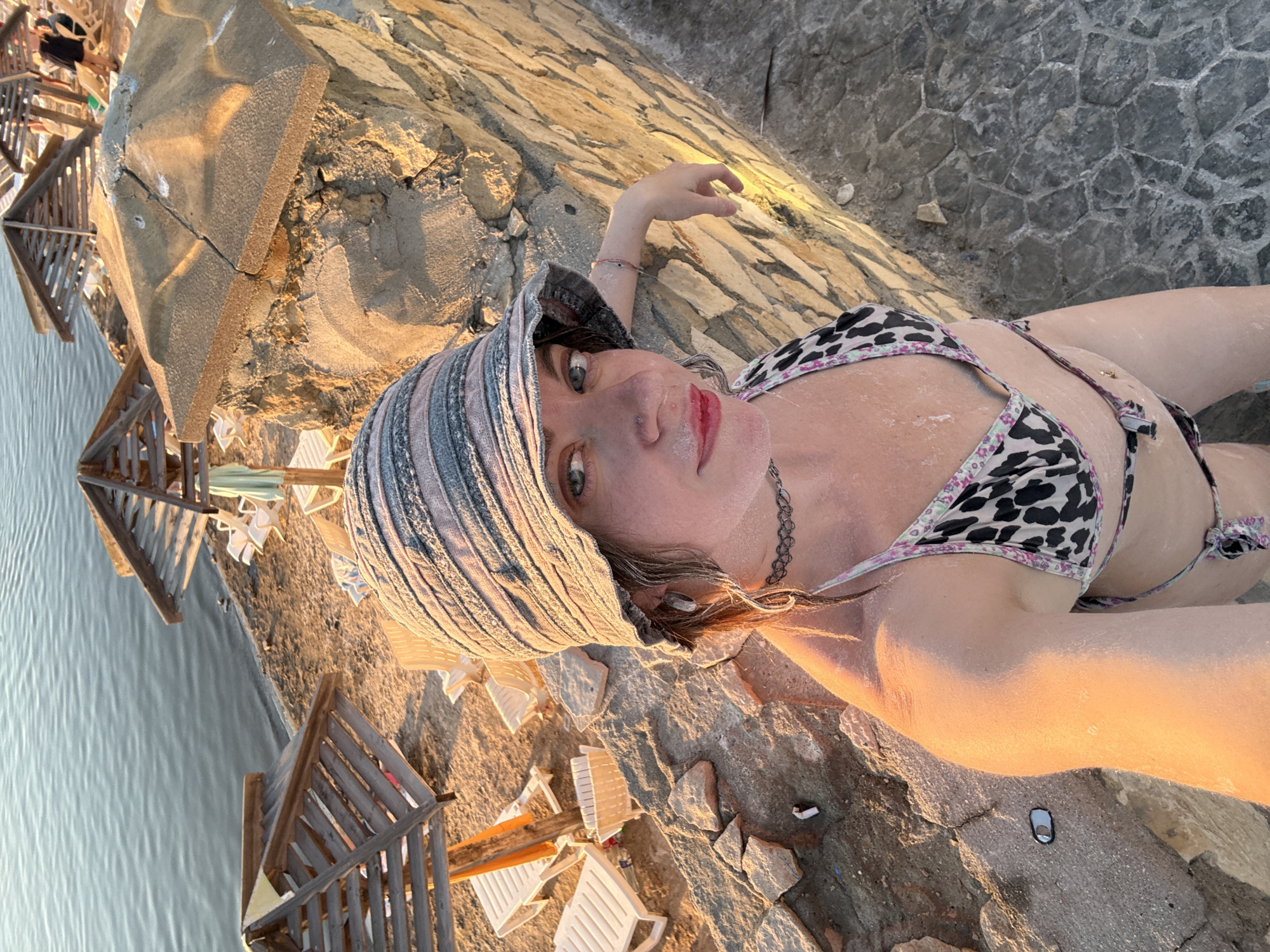 This media file shows a young woman lying down on a rocky surface next to a body of water. She is wearing a patterned bikini and a sun hat. Her skin appears to be dusted with a white substance, possibly salt or mud, consistent with a spa or therapeutic mud bath experience often found in Sol-Iletsk, Russia, known for its salt lakes and mud treatment centers.
The setting appears to be an outdoor area with several wooden structures that resemble small cabanas or changing rooms, and lounge chairs are visible near the water. The lighting suggests it is either sunrise or sunset, casting a warm, golden glow on the scene and the woman's skin. The water is calm, reflecting the soft light. The rocks are varied in size and texture, forming an uneven terrain.
The woman is looking towards the camera with a relaxed expression. Her hand is resting on the rocky ground, and her body is positioned to face the viewer. The presence of salt or mud on her skin and the overall environment strongly suggest she is participating in a wellness treatment associated with the mineral-rich waters and mud of the region. No visible text is present in the image.