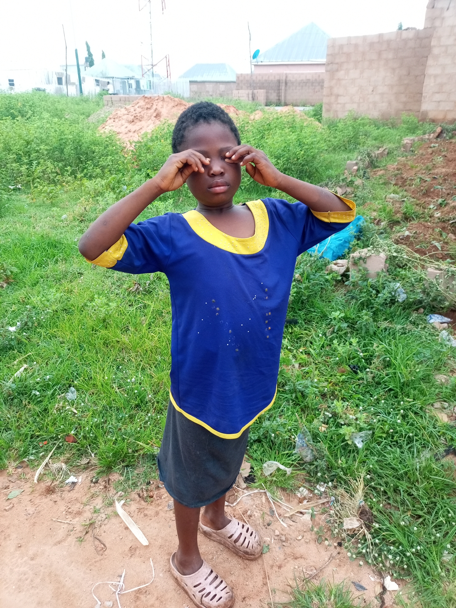 In Tempure, Nigeria, a young boy stands outside in a dusty, grassy area. He wears a blue t-shirt with yellow trim and dark shorts. His eyes are closed, and his hands are covering them, suggesting he may be experiencing discomfort or playing a game. The background shows a mix of green vegetation, dirt, and unfinished construction, with glimpses of buildings and a water tower in the distance. The scene appears to be a typical residential or undeveloped area on the outskirts of the city.