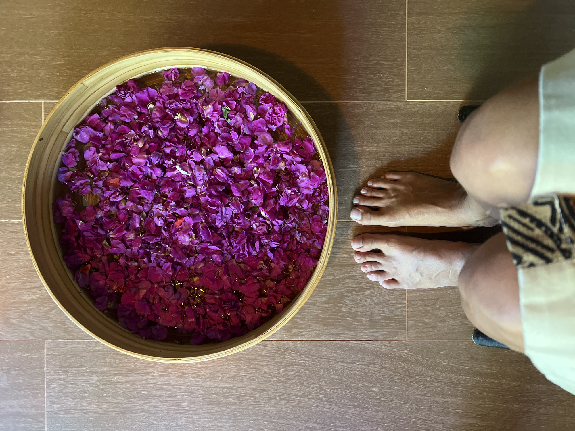 An overhead shot shows a person's bare feet positioned in front of a round, shallow bamboo bowl filled with water and vibrant purple flower petals. The bowl rests on a tiled floor composed of large, rectangular tiles in varying shades of brown and beige, arranged in a grid pattern. The scene appears to be an indoor setting, possibly a spa or a traditional Indonesian bathhouse, likely located in the Ubud District of Indonesia. The lighting is warm and somewhat dim, suggesting an ambient, relaxing atmosphere. The flower petals are abundant, creating a rich tapestry of color within the water. The person's feet are positioned centrally in the frame, with toes pointing forward towards the bowl, implying they are about to step into or have just stepped out of the floral foot bath.