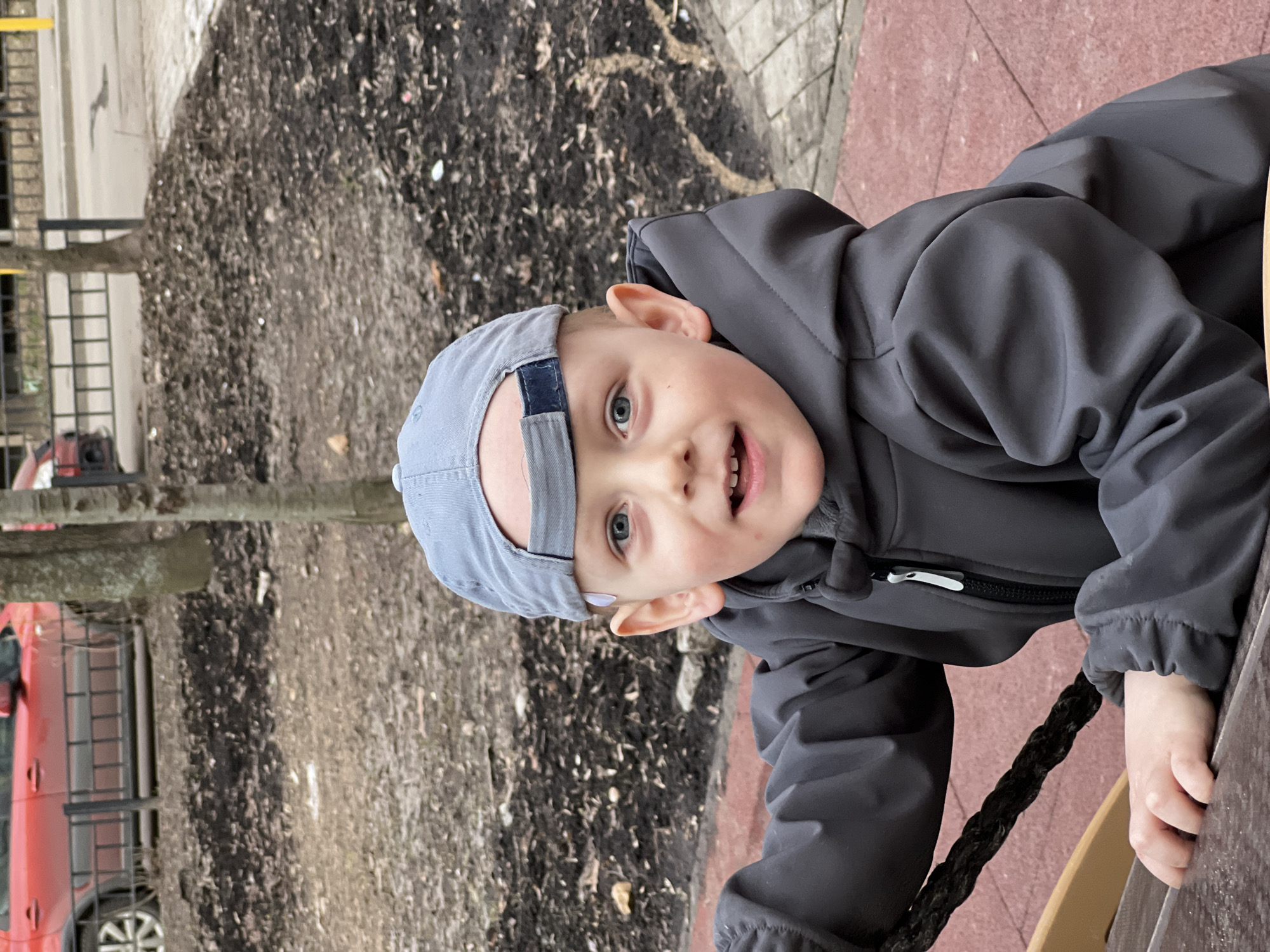 A young child wearing a grey hooded jacket and a light blue baseball cap is climbing a rope ladder in a park, seemingly located in Mytishchi, Russia. A section of park ground covered in dark mulch can be seen in the background, along with a red vehicle in the distance. The surface below the ladder appears to be a dark pink paved area. The child is looking at the camera with a slight smile.