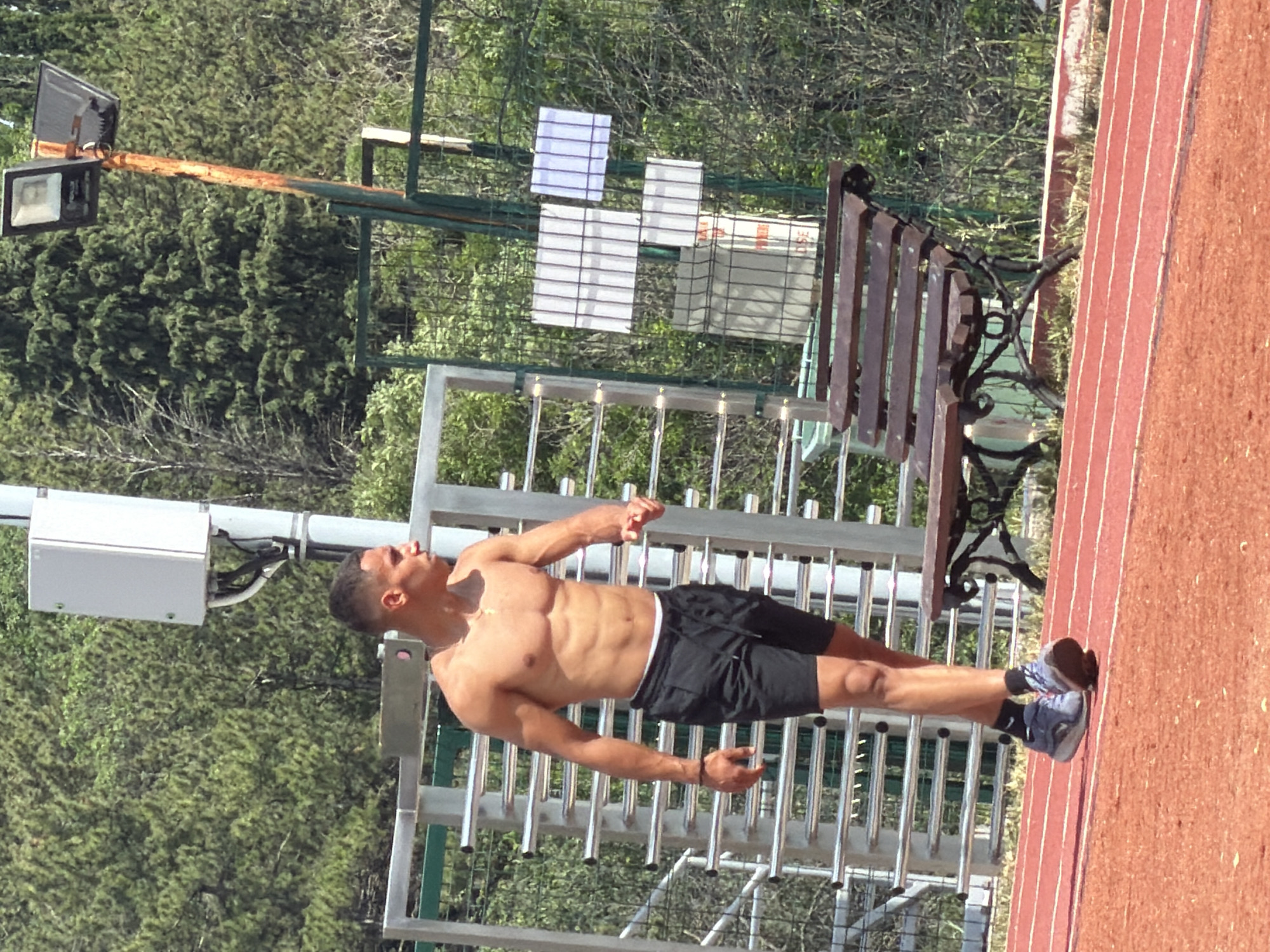 The image depicts a muscular man posing at an outdoor athletic facility in Stara Zagora, Bulgaria, under bright daylight. The shirtless man, with a toned physique and dark hair, is wearing black athletic shorts, dark socks, and grey and blue athletic shoes. He stands upright with his arms slightly extended, facing forward, showcasing his condition. He also wears a thin necklace.
In the immediate foreground on the right side of the image, a red running track with white lane lines is prominently visible. The man is positioned next to a silver-colored metal structure, likely outdoor calisthenics or workout equipment, which features multiple horizontal bars.
The background is dominated by lush green trees and dense foliage. A fence runs horizontally across the middle ground, separating the workout area from the treeline. Attached to this fence are several white rectangular boards or signs, some with horizontal markings, and what appears to be a cardboard box. In the upper left background, a tall pole supports two black floodlights. Closer to the man, on the left side, a white rectangular utility box is attached to another pole or structure.
Further in the background, behind the track, a wooden slatted park bench with an ornate black metal frame is partially visible. The clear lighting suggests a sunny day. There is no discernible text visible in the image. The overall scene conveys an active, outdoor fitness environment.