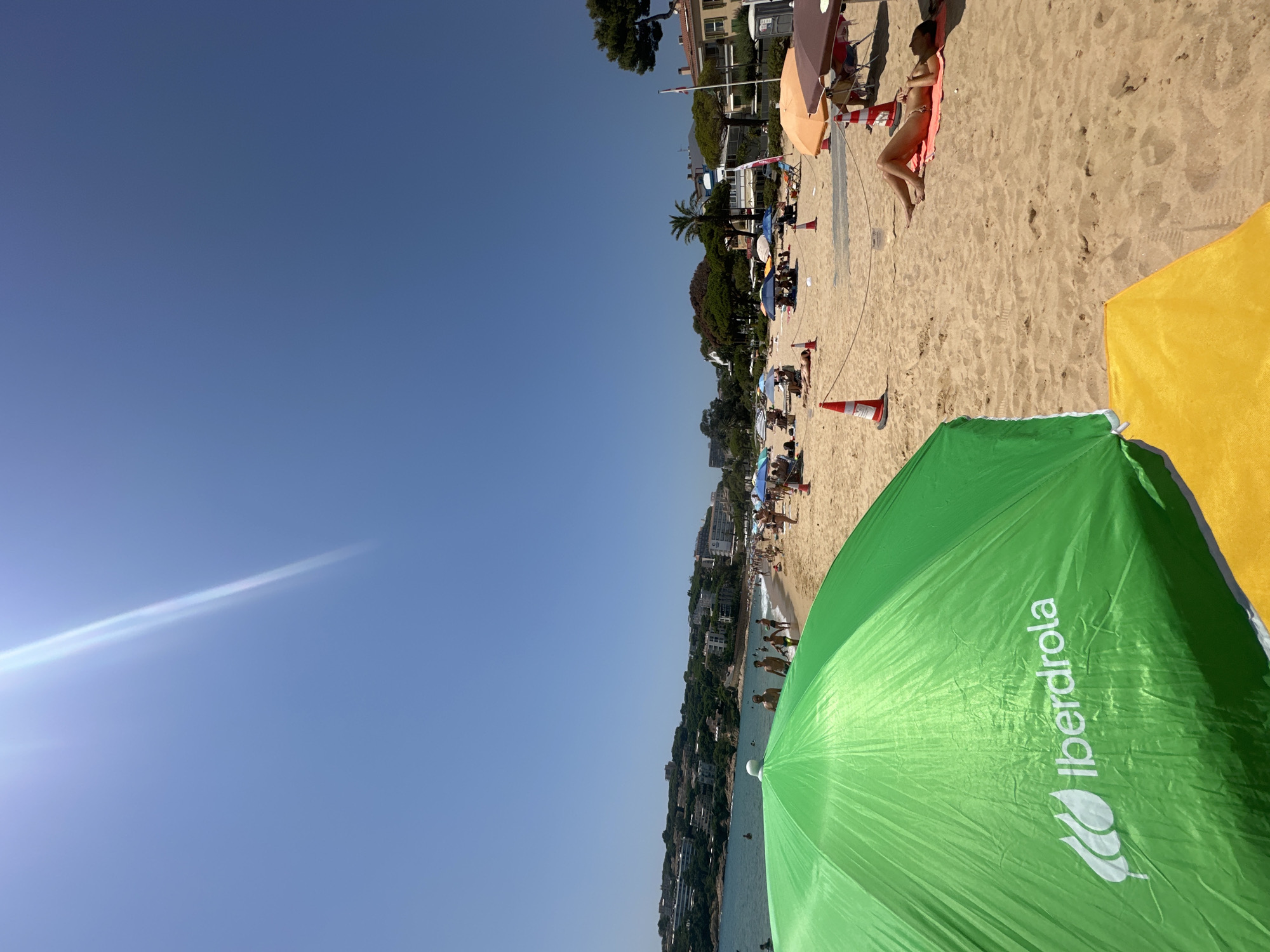This is a full-frame, eye-level, outdoor, daytime photograph of a crowded beach on a sunny day in Sant Feliu de Guíxols, Spain. The foreground is dominated by a large, bright green beach umbrella with the white Iberdrola logo visible. A portion of a yellow beach towel or umbrella is also visible in the lower right corner. The sand on the beach is light brown and appears dry.
Further back, numerous people are scattered across the beach, some under umbrellas, others sunbathing, and many are walking along the shoreline. The water of the Mediterranean Sea is a clear blue and several individuals are visible in the water. Along the coastline, a dense collection of buildings, hotels, and residential structures are built on the hillsides overlooking the beach. Vegetation, including trees and palm trees, is also present on the hills. The sky is a clear, bright blue with a prominent sun flare streaking diagonally from the upper left to the center of the frame. The overall scene depicts a lively and popular summer beach environment.