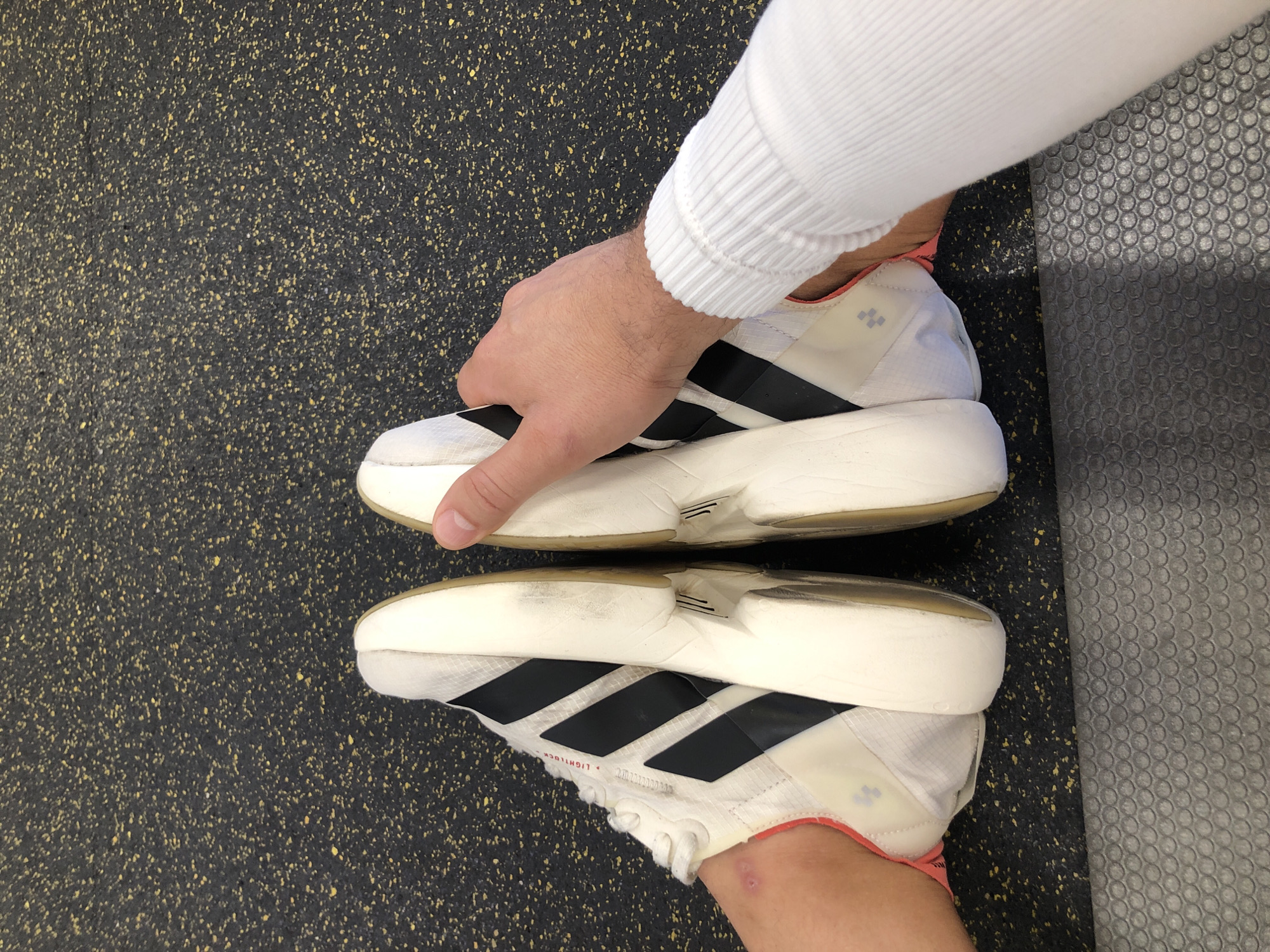 The image shows a close-up view of two white athletic shoes with black stripes. The shoes are placed on a speckled dark gray floor, possibly a gym or sports facility, with some yellow flecks visible. A person's hand wearing a white ribbed sleeve is holding the top shoe, suggesting they are either putting on or taking off the shoes. The bottom shoe is on the person's foot, also partially visible. The lighting is bright, indicating an indoor setting. The background includes a grey textured mat with a circular pattern. There is no indication of the time of day or weather conditions. There is no visible text in the image.