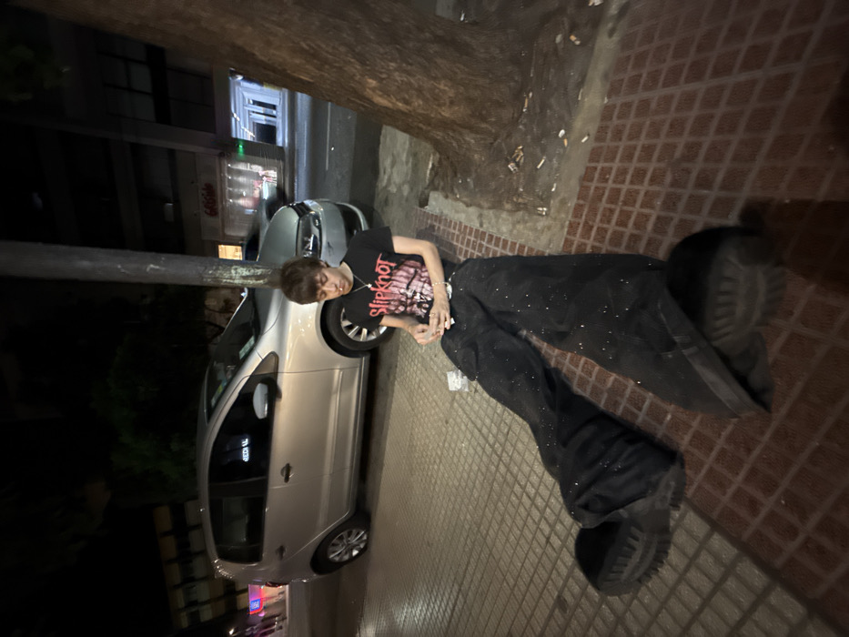 A person is lying on a brick sidewalk, possibly unconscious. They are wearing a black t-shirt with a Slipknot logo, black pants, and dark shoes. A car is parked nearby. A tree is next to the person, with numerous cigarette butts scattered on the sidewalk around its base. The scene appears to be at night, with streetlights illuminating the area. The image is tilted.
