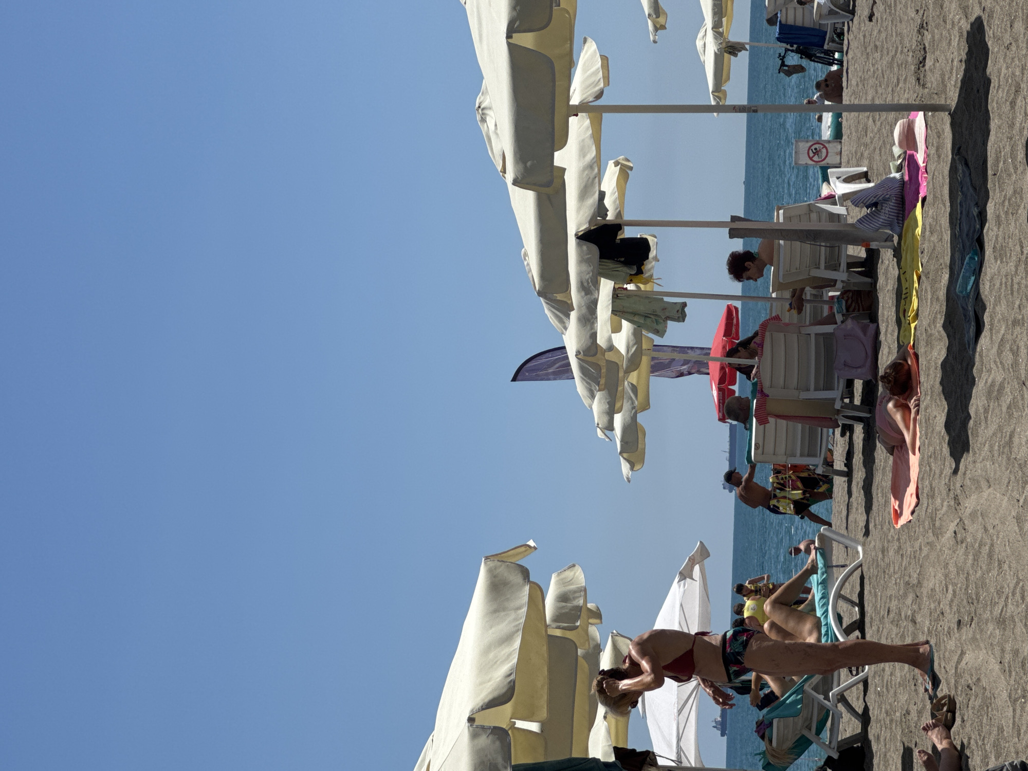 The image depicts a crowded beach scene with multiple beach umbrellas and sunbathers. In the foreground, a woman in a red bikini stands with her hand on her head. Several people are lying on lounge chairs or towels under umbrellas. In the background, the ocean is visible with a few ships in the distance. The sky is clear and blue. The location is not specified as Burgas, Bulgaria.