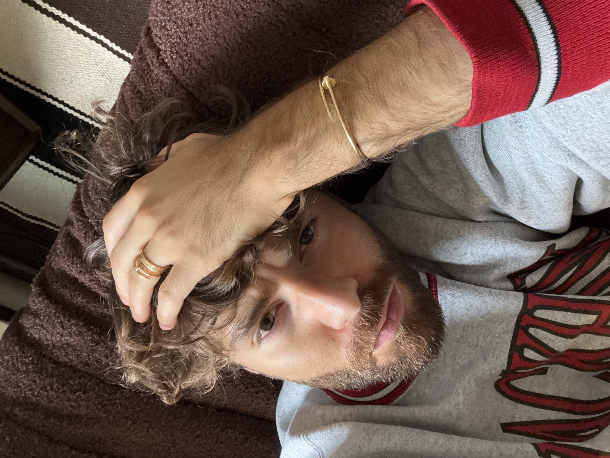 A man with curly brown hair, a trimmed beard, and wearing a gray and red sweatshirt is lying down, possibly on a couch. He has a gold ring on his finger and a gold bracelet on his wrist. The background includes a brown fabric and striped fabric. The location is not discernible as Montreal, Canada, or any other specific location.