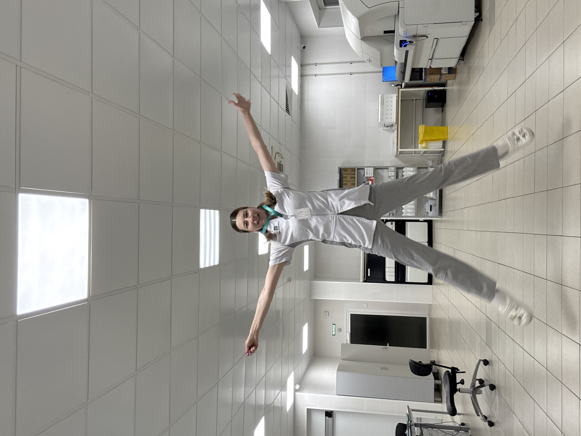 A young woman in a white lab coat and gray pants is jumping in what appears to be a laboratory setting. She is in mid-air with her arms and legs spread out in a star shape. She is holding a small, purple object in her right hand. The room has a tiled floor and a suspended ceiling with fluorescent lights. There are various pieces of laboratory equipment visible in the background, including a large white machine with a blue computer screen, shelves filled with medical supplies, and a yellow trash can. The woman has a lanyard around her neck with an ID badge. The environment is clean and organized, typical of a medical or research facility. There is no indication of this location being in Moscow, Russia, or any specific city.