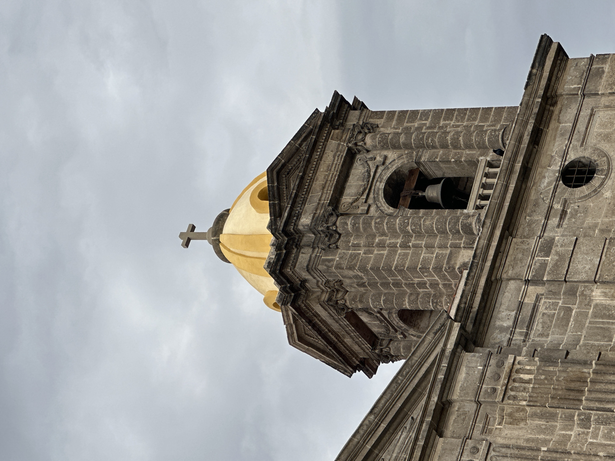 The image shows a close-up, upward view of a church tower with a golden dome in Mexico City, Mexico. The tower is constructed of gray stone and features detailed stonework, including arches, columns, and decorative moldings. A bell is visible in one of the tower's arched openings. Atop the golden dome is a simple cross. The sky is overcast and gray.