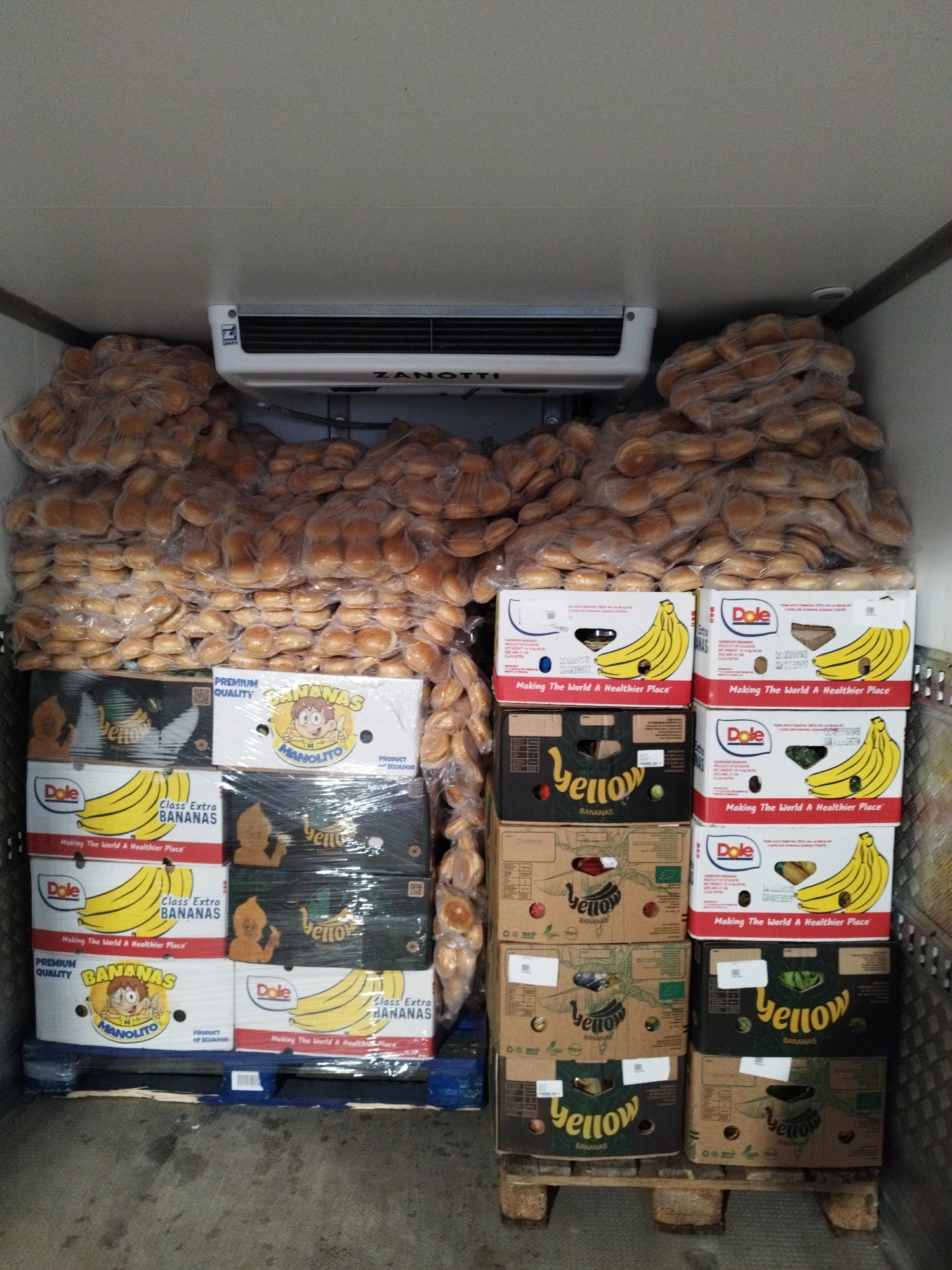 The image shows the interior of a refrigerated truck. The truck is filled with stacks of boxes and bags of food items. In the upper section of the truck, there is a large white refrigeration unit with the brand name "ZANOTTI" visible. Below the refrigeration unit, the walls of the truck are white.
The majority of the space is occupied by large, clear plastic bags filled with numerous small, round, baked goods, likely hamburger or sandwich buns. These bags are stacked high, filling the rear and side portions of the truck.
In the foreground, there are stacks of cardboard boxes containing bananas. Some of these boxes have images of bananas and branding from "Dole" and "Manolito". There are also some boxes with a green and black design featuring the word "yellow" and stylized bananas. The boxes are stacked on blue and wooden pallets.
The floor of the truck appears to be a rough, possibly concrete or tiled surface, with some dirt and debris. The overall impression is one of a commercial food storage or transport environment. The location is Cernica, Romania. There are no people visible in the image.