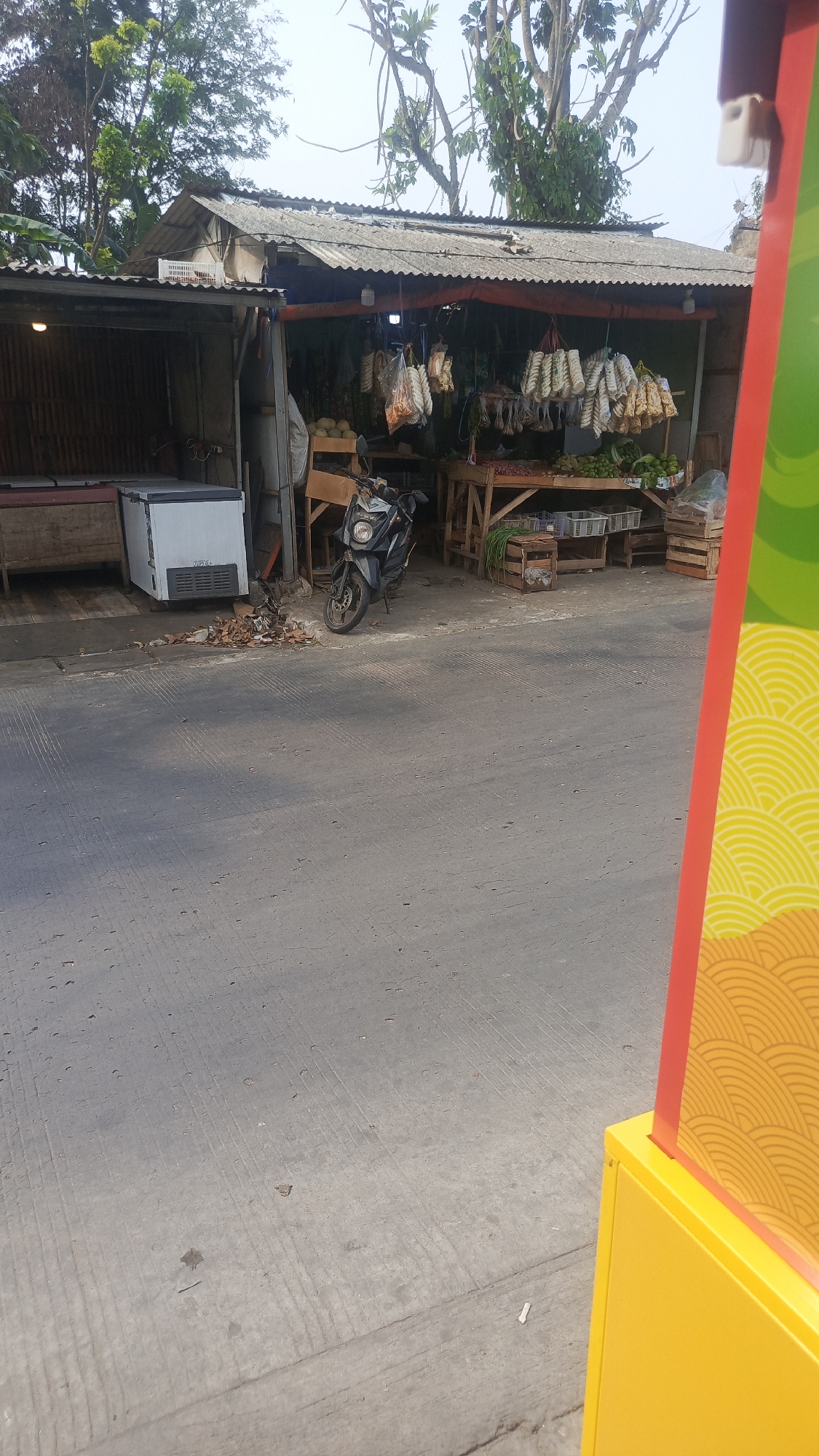 This is a photograph of an outdoor market stall in Parung Panjang, Indonesia, captured from a low angle. The stall features a corrugated metal roof supported by wooden posts and covered with an orange tarp. Rows of bagged goods, possibly snacks or dried items, hang from the roof. A wooden table displays various fresh produce, including what appear to be green fruits or vegetables. Two white plastic crates are visible beneath the table, and a motorcycle is parked to the left of the stall. To the left of the stall, a white chest freezer sits on a wooden platform. The ground is paved with asphalt, showing tire marks and scattered debris, including dried leaves. In the foreground, a brightly colored structure with red and yellow panels, adorned with a yellow wave pattern, partially frames the right side of the image. The background consists of trees and a cloudy sky. No people are visible in the scene.