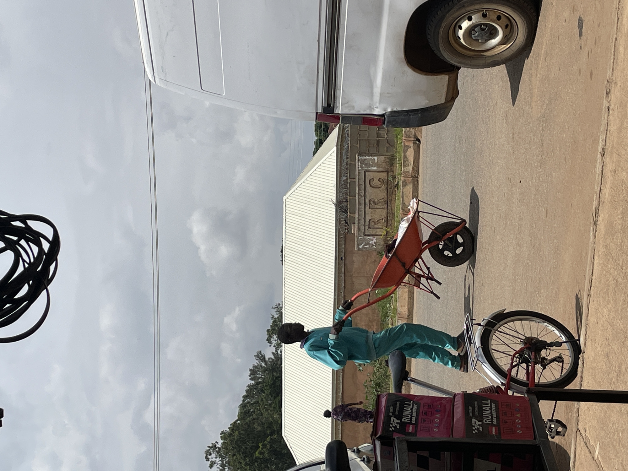 A medium shot captured on a cloudy day in Bukuru, Nigeria, shows a man in a teal tracksuit pushing an orange wheelbarrow along a paved road. He is walking from left to right across the frame. In the background, a woman in a patterned dress is visible walking away from the viewer. A white van is parked on the right side of the image, obscuring part of the background. The road is lined with a low wall and vegetation. In the foreground, a bicycle with a cargo rack loaded with what appear to be car batteries is positioned on the left. Overhead, a thick bundle of black cables is suspended.