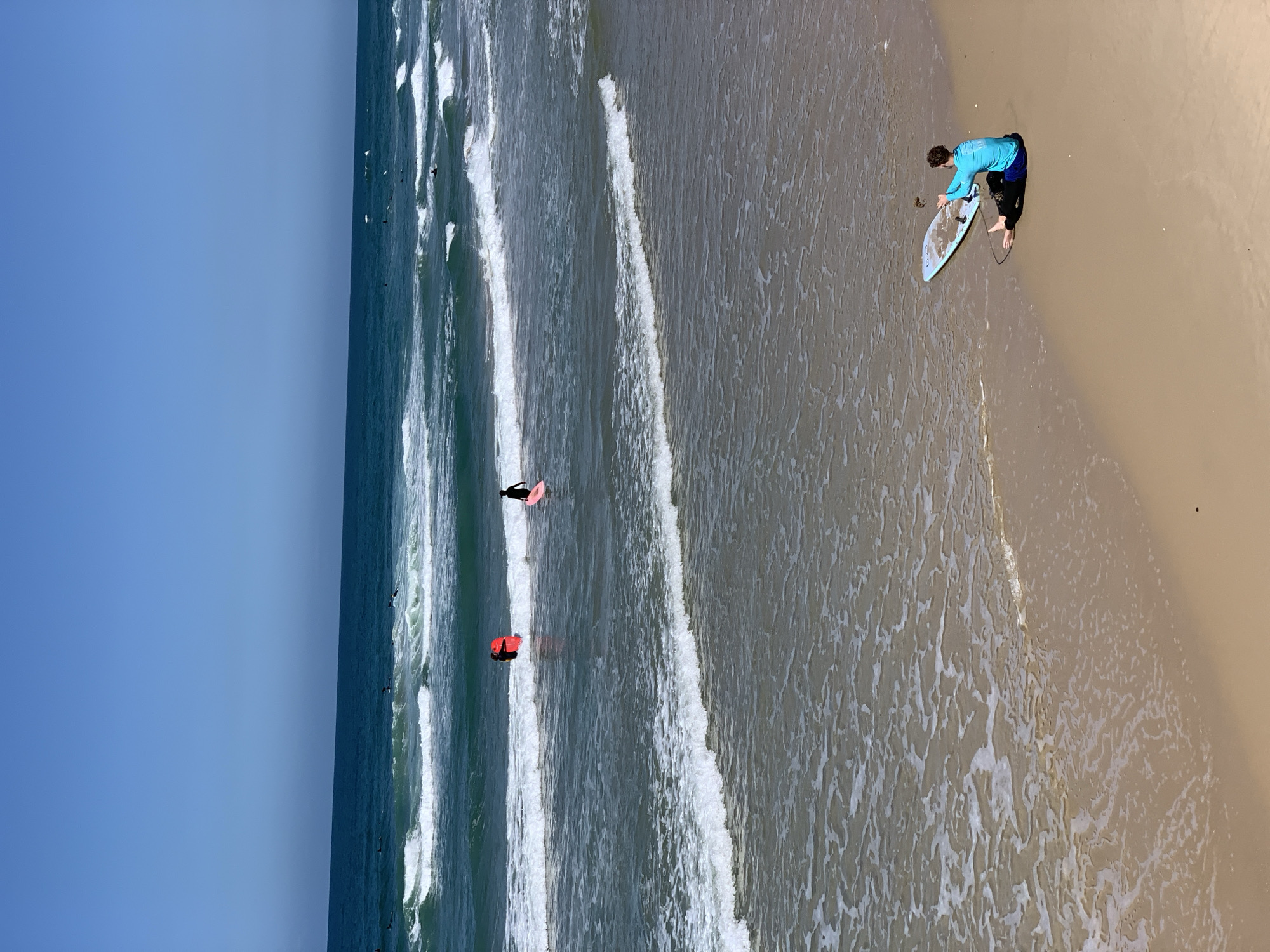 The image shows a beach scene in Tel Aviv, Israel. The view is from above, oriented vertically. A young male stands on the sand at the water's edge, holding a light blue skimboard. He wears a bright blue rash guard and dark-colored shorts or wetsuit bottoms.
In the ocean, there are several surfers and bodyboarders, some visible amidst breaking waves. One bodyboarder has a bright red board, and another has a pink one. The sea transitions from a lighter, foamy area near the shore to a deeper blue further out. The sky is clear and bright blue. The sand is a light tan color, with patterns created by the receding waves.