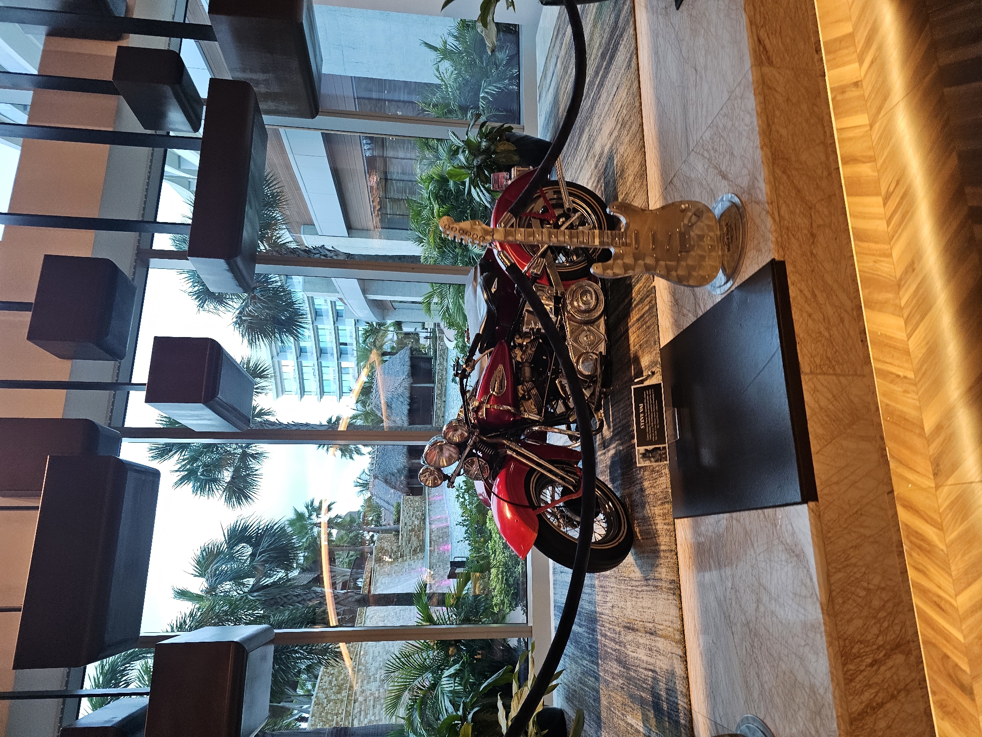 The image depicts an indoor display featuring a red motorcycle and a clear electric guitar. The backdrop is a large window offering a view of a tropical landscape, including palm trees and buildings, likely within a resort or entertainment complex. To the left, there's a decorative lighting fixture with rectangular elements. A sign reading 'Steve Vai' is visible next to the motorcycle. The scene is set at the Seminole Hard Rock Hotel & Casino in Hollywood, Seminole Reservation, Florida, United States of America.