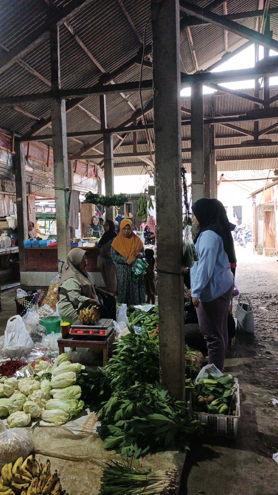 This image depicts an outdoor market scene in Cibenda, Indonesia, characterized by a rustic, open-air structure with a corrugated metal roof supported by concrete pillars. The market stalls are laden with a variety of fresh produce, including leafy green vegetables, cabbage, cauliflower, bananas, and chilies. Several individuals, predominantly women wearing headscarves, are present, engaged in activities typical of a marketplace, such as selling, purchasing, or interacting with each other. One woman is seen using a digital scale to weigh produce. The ground appears to be unpaved, with loose dirt and debris. The overall atmosphere suggests a bustling local market where fresh goods are exchanged.