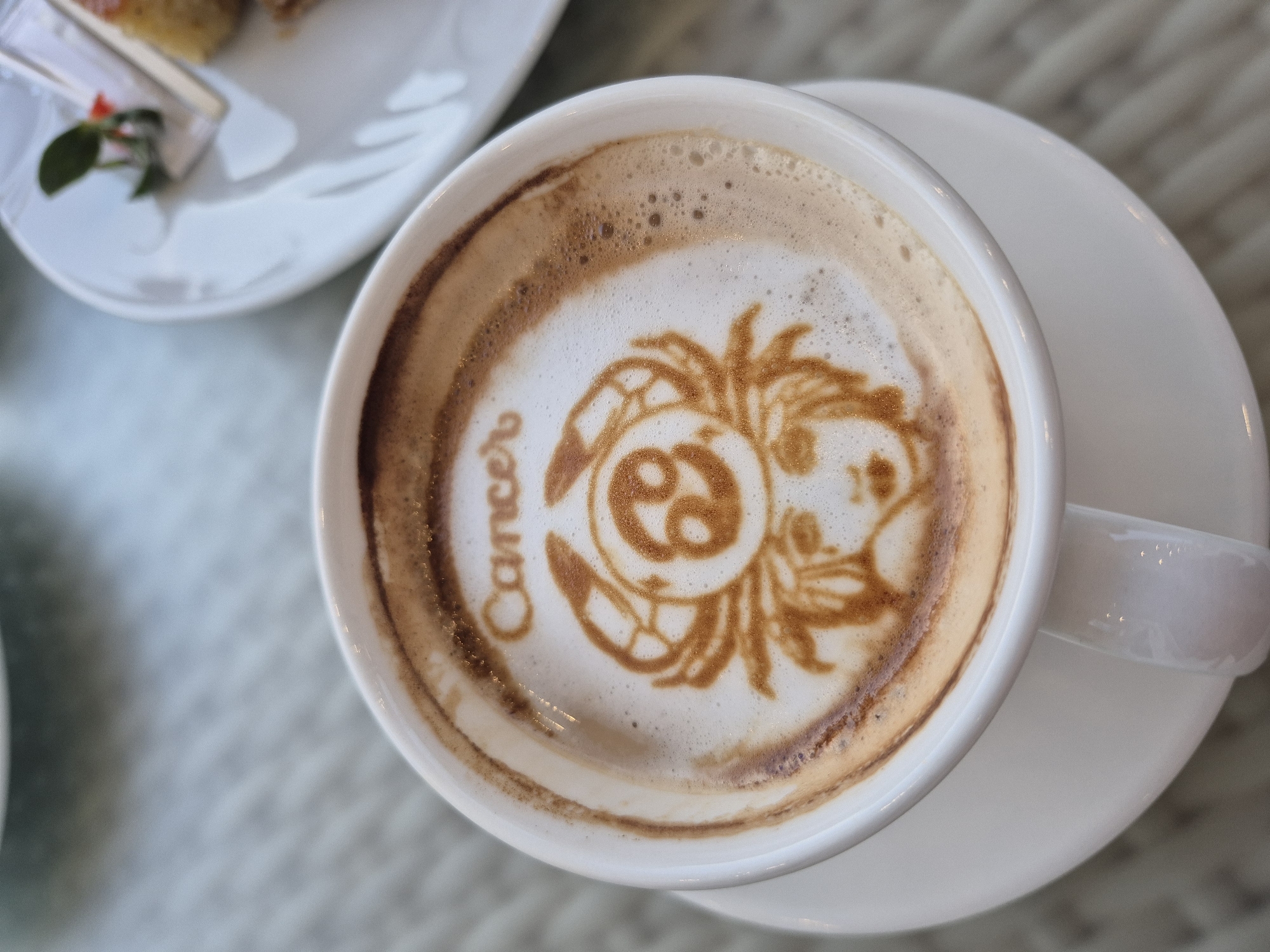 Here's a description of the image:
A top-down, close-up shot shows a white ceramic cup filled with latte art. The art depicts the astrological sign Cancer, complete with the word "Cancer" written along one side. The latte art is composed of dark brown foam. The cup sits on a white saucer. A white plate with a small dessert item is partially visible in the upper left corner of the frame. The background appears to be a light-colored woven surface. The location and city are not discernible from the provided image.