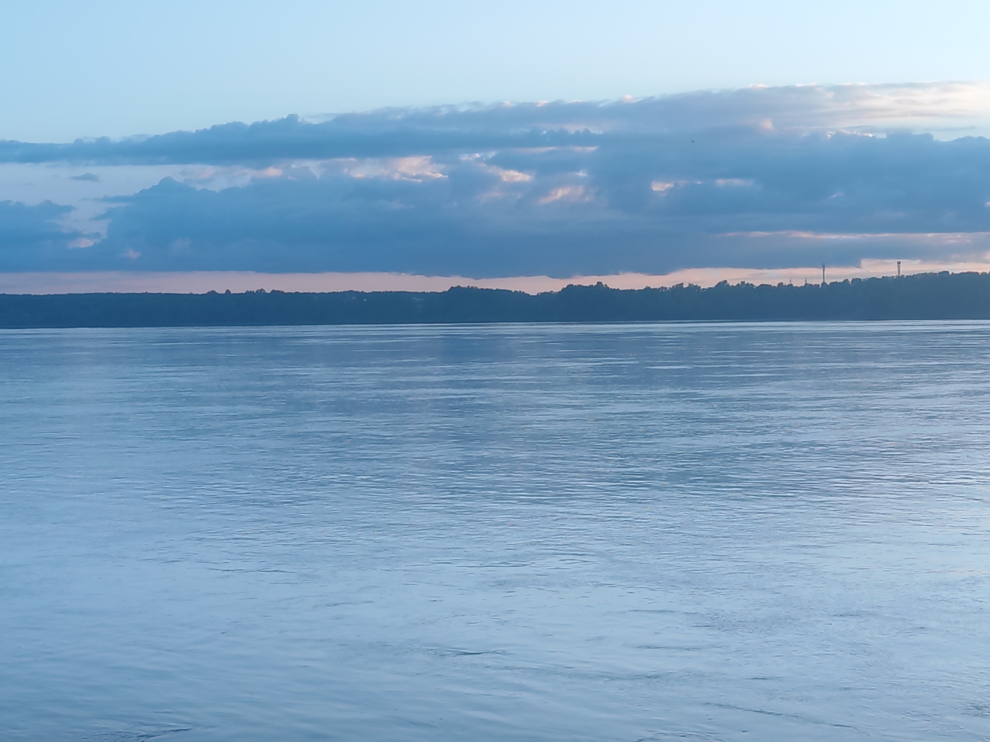 The image presents a serene landscape dominated by a wide body of water under a cloudy sky, likely captured during either sunrise or sunset given the soft, diffused light. The water, which appears to be a large river or lake, occupies the majority of the lower two-thirds of the frame, exhibiting subtle ripples and a cool blue-gray hue.
A distant, low-lying landmass, densely covered with trees, forms a dark, silhouetted horizon line across the middle of the image. Above this tree line, the sky is filled with multiple layers of clouds, predominantly in various shades of blue and gray. Hints of soft pink and orange light peek through or illuminate the edges of some clouds, particularly closer to the horizon, reinforcing the sense of twilight. On the far right of the distant horizon, two faint, tall, slender structures, possibly communication towers, are barely visible against the sky.
The scene conveys a sense of tranquility and vastness. There are no visible people or explicit activities taking place, and no discernable text is present. The overall atmosphere is peaceful and natural.
