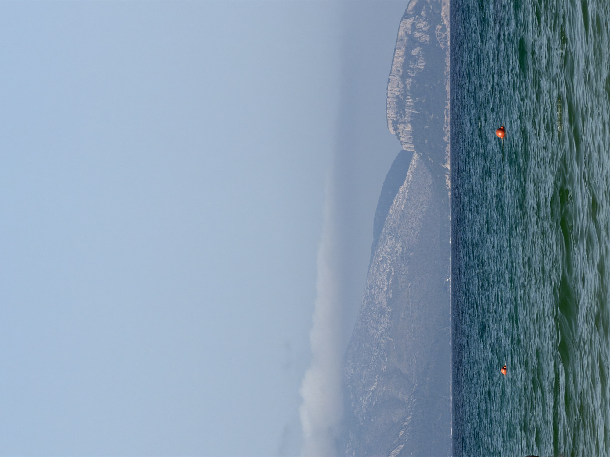 The image depicts a calm sea with rolling waves under a clear, pale blue sky. In the distance, a range of rugged mountains rises, some with patches of vegetation and rocky outcrops. A thin layer of clouds hovers over the peaks of the mountains. Several sailboats are visible on the horizon. Two bright orange buoys float in the turquoise water. The foreground shows the green, choppy surface of the sea. The scene appears to be a coastal landscape, likely near Olbia, Italy, given the characteristic Mediterranean mountainous coastline and clear blue waters.
