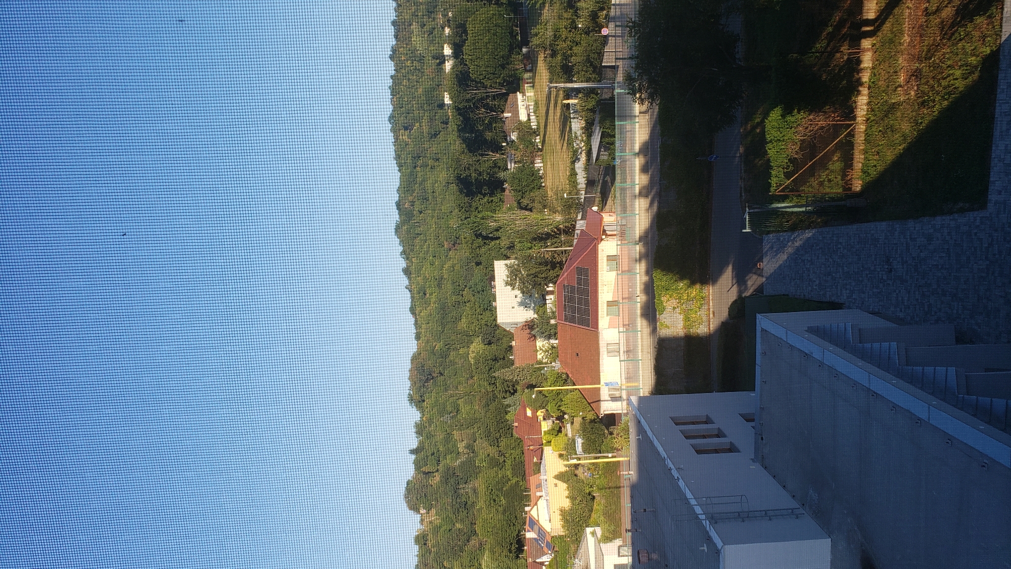 The image shows a view from a window with a screen, looking out over a residential area with a wooded hill in the background. In the foreground, there are the rooftops of modern buildings. Further back, there are houses with red tiled roofs, some with solar panels. A street and a bridge are visible. The sky is clear and blue, with a few small dark specks, possibly birds or insects. The scene appears to be a suburban or semi-rural environment. The image does not contain any specific identifiable landmarks or signs that would allow for precise location within Prešov, Slovakia, beyond it being a general view of a town or city in that region.