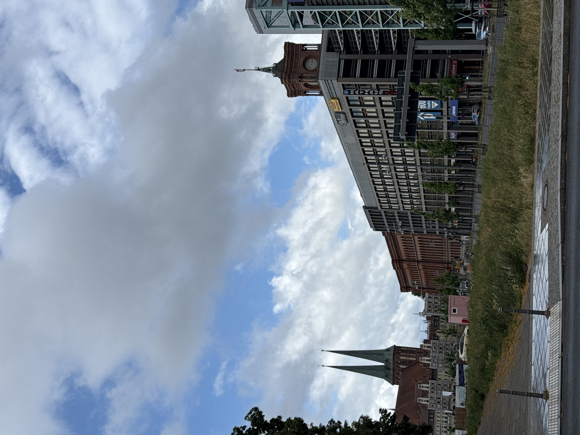 The image shows a city scene in Berlin, Germany, likely on a partly cloudy day. On the left, a historic church with two tall green spires is visible, with older buildings behind it. To the right of the church, there is a modern, multi-story building with a red brick facade. Further to the right, a large, contemporary building with a gray facade stretches across the frame, featuring numerous windows and balconies. A distinctive red brick tower with a clock is attached to this building. Several trees line the sidewalk in front of the contemporary buildings. In the foreground, a paved walkway with tactile paving runs alongside a street. A patch of tall, dry grass is situated between the walkway and the buildings. The sky is filled with a mix of white and gray clouds.