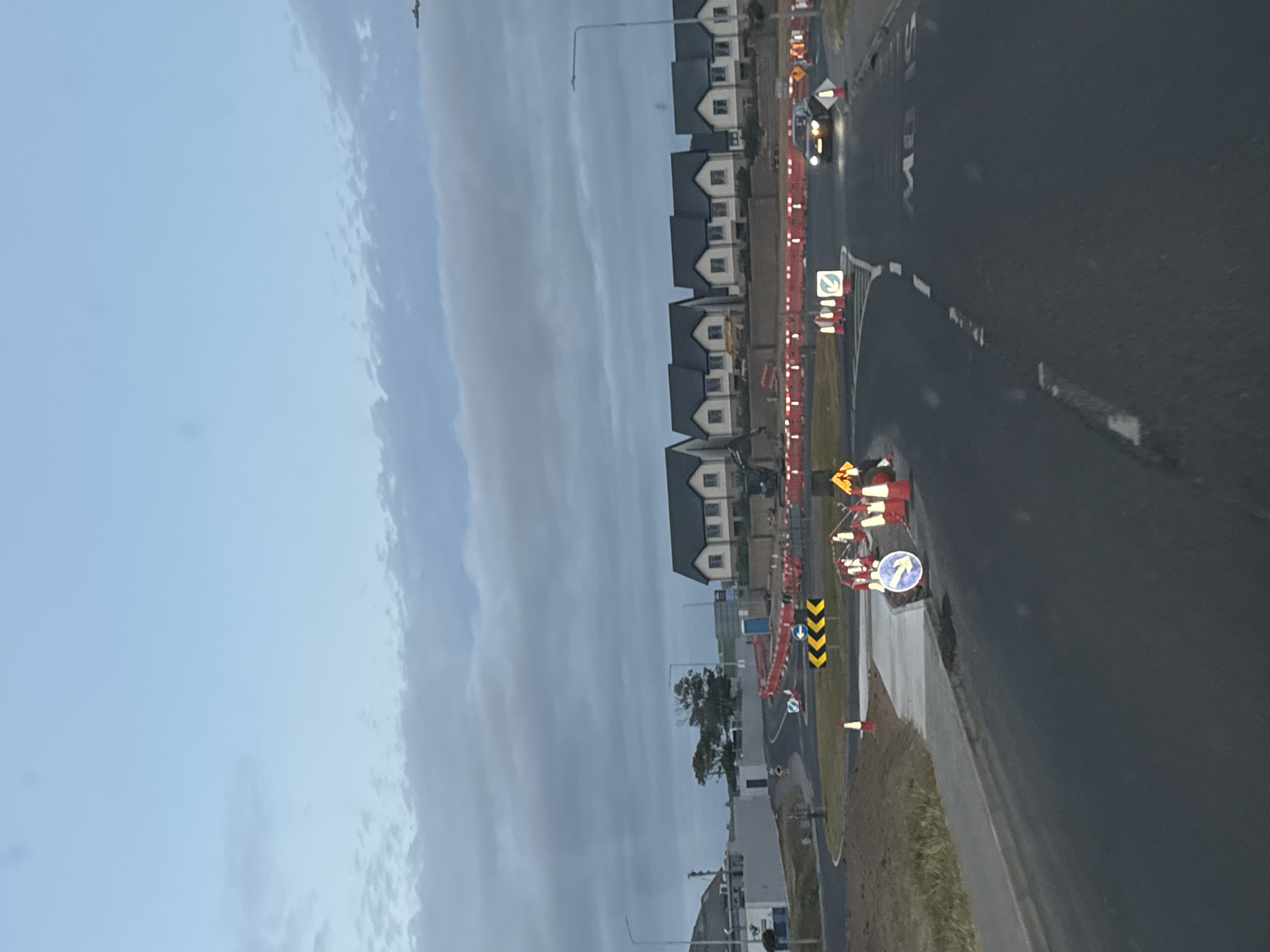 The image depicts a construction site with numerous orange barriers and traffic cones along a road in Tramore, Ireland. A car is visible on the road, and several houses are situated in the background. The sky is overcast with clouds. The scene suggests ongoing roadwork or development in a residential area.