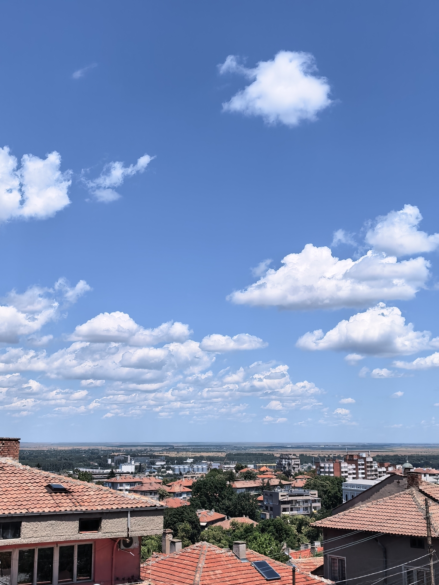 The image is a high-angle, full shot of a city skyline under a bright blue sky with scattered white clouds. In the foreground, several rooftops with traditional orange tiles are visible, some with solar panels. The middle ground shows a densely populated urban area with buildings of varying architectural styles, including apartment blocks and smaller houses, interspersed with trees. Beyond the city, a flat landscape stretches to the horizon, meeting the sky. The distant landscape appears to be a mix of agricultural fields and industrial structures, including silos. The scene suggests a sunny day in a city, possibly Svishtov, Bulgaria, given the presence of a dome on one of the buildings which could be a church. There are no people visible in the image.