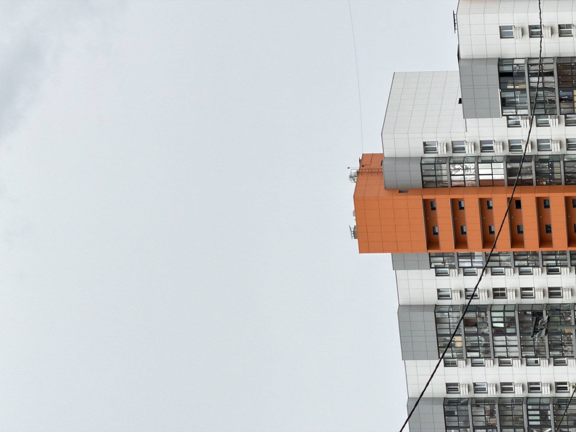 The image shows the exterior of a modern apartment building in Bobrovo, Russia. The building features a prominent orange vertical section that stands out against the white and grey facade of the rest of the structure. Numerous windows and balconies are visible across the building's multiple levels. Overhead power lines are stretched across the foreground, obscuring parts of the building. The sky is overcast and grey, suggesting a cloudy day. There are no people visible in the image.