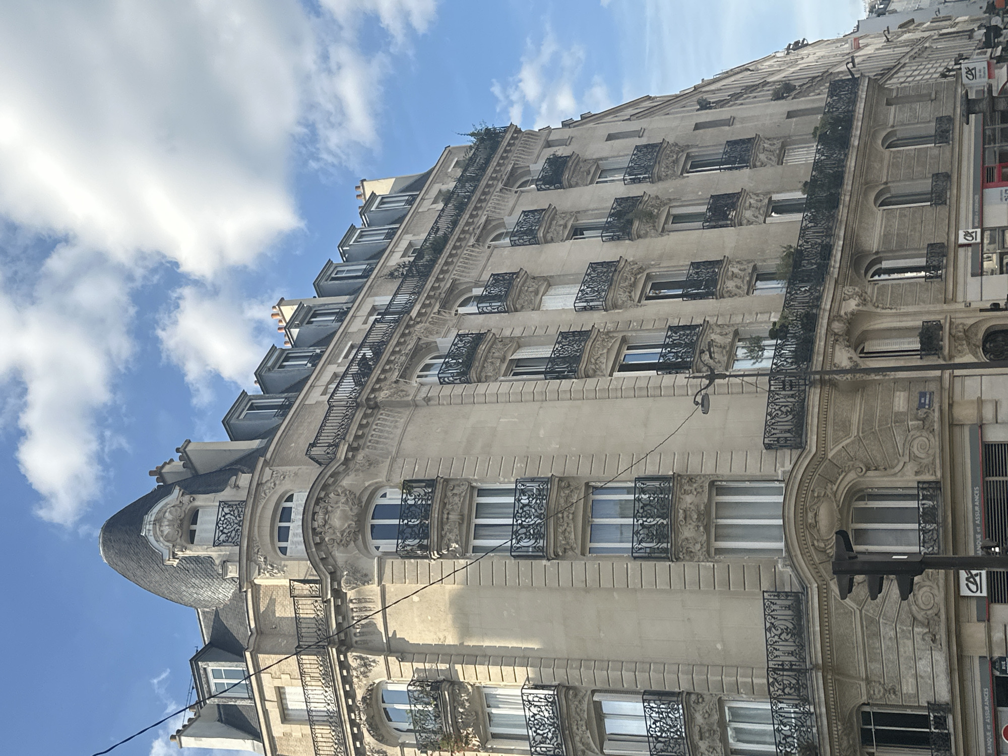 An eye-level, medium shot captures a Haussmannian building in Paris, France, under a partly cloudy blue sky. The building, a typical Parisian architectural style, features a stone facade with ornate balconies and mansard roofs. Sunlight illuminates the right side of the building, creating shadows on the left.
The foreground shows a street-level view of the building's lower floors. A traffic light with three lights is positioned in the center-left of the frame. The building's ground floor appears to be commercial, with signage for "CA BANQUE et ASSURANCES" visible.
The building itself is detailed and grand, with multiple stories showcasing a repeating pattern of windows and balconies. The balconies are decorated with intricate black wrought iron railings. The mansard roofs are covered in dark slate, with dormer windows protruding from them. A distinctive rounded turret with a pointed, slate-covered roof sits atop the corner of the building.
The sky is a vibrant blue with scattered white, puffy clouds. The overall impression is one of a classic Parisian street scene, bathed in natural light. There are no people clearly visible in the image, and no specific actions or interactions are depicted. The scene is static and focuses solely on the architecture and atmosphere of the location.