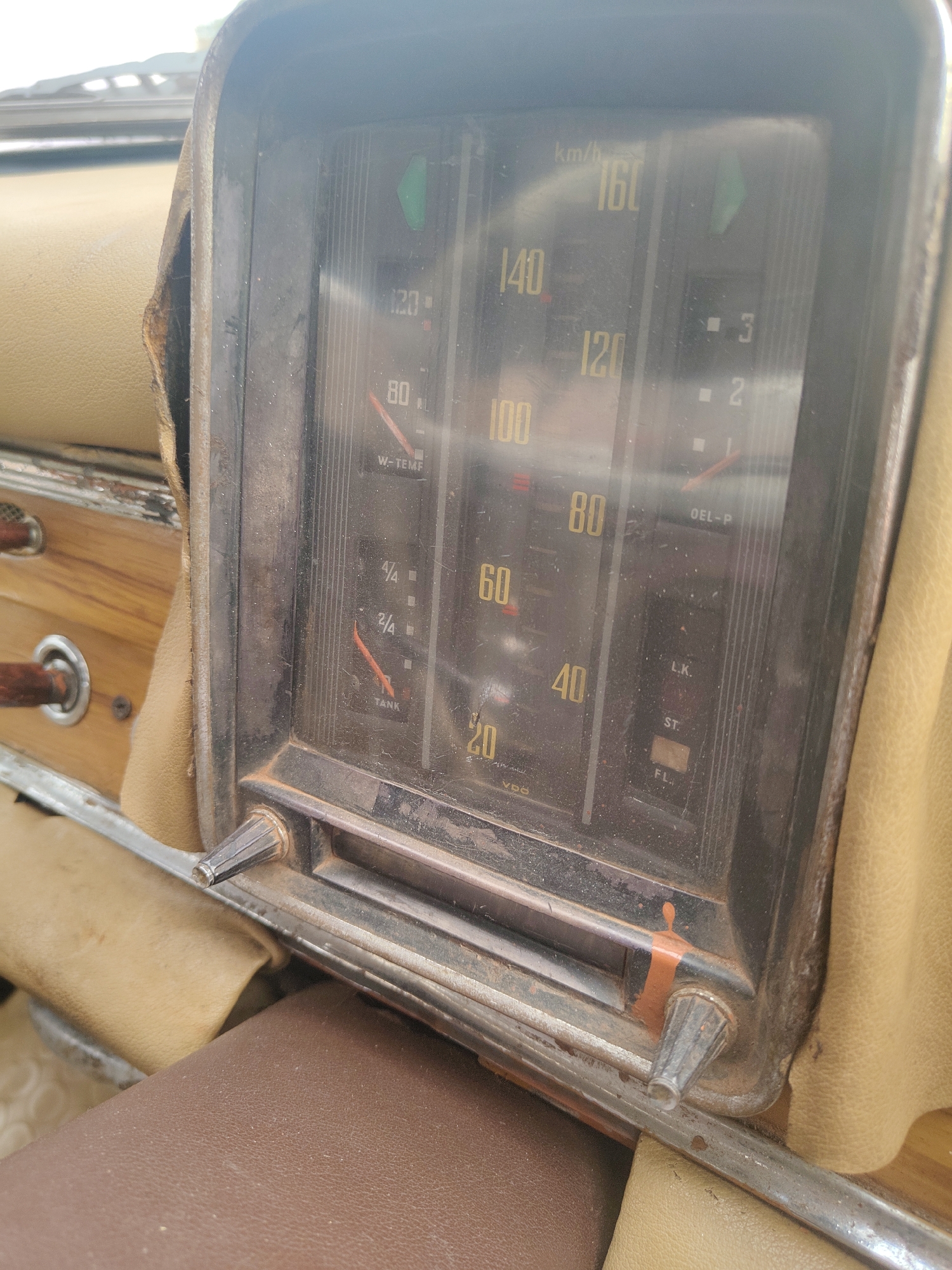 The image displays the worn dashboard of an old car, likely shot from a passenger's perspective in Kalamassery, India. The central subject is a rectangular instrument cluster, appearing vintage, dirty, and somewhat rusty around its metallic frame. The surrounding interior upholstery, in shades of tan and brown, shows signs of age and tearing.
The instrument cluster features several analog gauges behind a dusty, reflective glass. The main speedometer is on the left, marked in km/h, with visible numbers 20, 40, 60, 80, 100, 120, 140, and 160. Its needle rests at 0 km/h. Below the speedometer is a "W-TEMP" (water temperature) gauge with a low reading, and a "TANK" (fuel) gauge indicating a low fuel level, around 1/4 full or less, with markings "4/4" and "2/4". The bottom left corner of the cluster shows the manufacturer's name, "VDO".
To the right, additional indicators are visible. There's an "OEL-P" (oil pressure) gauge, also showing a low reading, and small square indicators labeled 1, 2, and 3. Further down, text labels "L.K.", "ST.", and "FL." are present, possibly for warning lights or other functions. A green indicator light is visible in the upper right section of the cluster. Two pointed knobs, possibly for resetting the trip meter or adjusting settings, protrude from the bottom of the instrument panel, with one showing a streak of orange paint or rust.
The overall scene conveys an impression of an aged vehicle, well-used and not in pristine condition, possibly stationary due to the zero speed reading and low gauge levels. The bright lighting suggests it is daytime.
