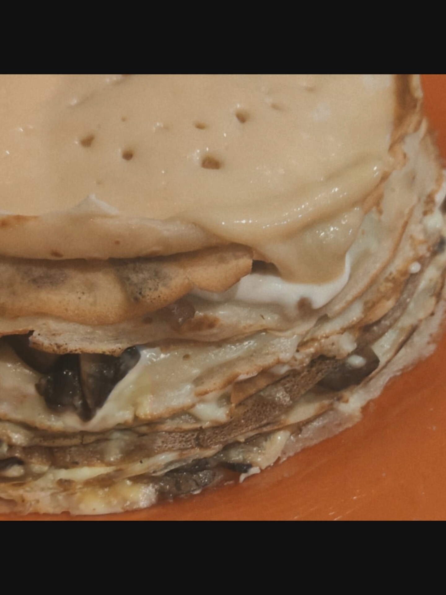 A close-up, overhead shot shows a stack of thin, layered crepes, possibly a savory crepe cake, on an orange plate. The crepes are golden brown, with some slightly darker edges, and are layered with a creamy filling that appears to be a light-colored sauce, possibly cream or béchamel, and visible dark pieces, likely mushrooms. The top crepe is slightly uneven and appears to be coated in the same creamy filling. The background is dark, with the bottom of the image showing the edge of the orange plate. There is no indication of the location in Saint-Août, France.