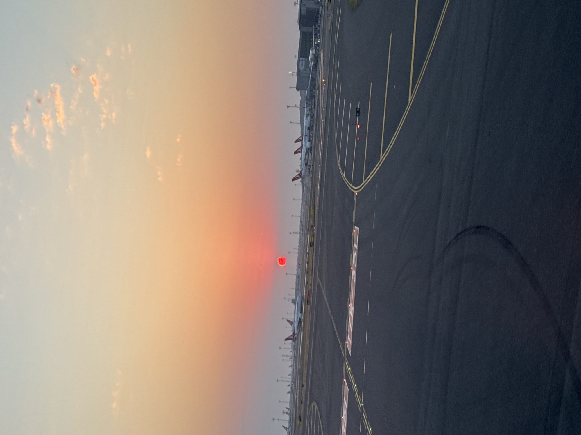 The image depicts an airport tarmac at sunset in Arnavutköy, Turkey. The sky is a gradient of warm colors, with hues of orange and pink. The sun, a bright red orb, is partially obscured by atmospheric haze and appears low on the horizon, casting a reddish glow across the scene. Several airplanes, identifiable by their white fuselages and red tail fins, are parked or taxiing on the runways and at gates. The tarmac is marked with yellow and white lines, and various lights illuminate the ground. The perspective suggests the photo was taken from a window of a terminal building or another aircraft. The overall impression is one of an active airport during the transition from day to night.