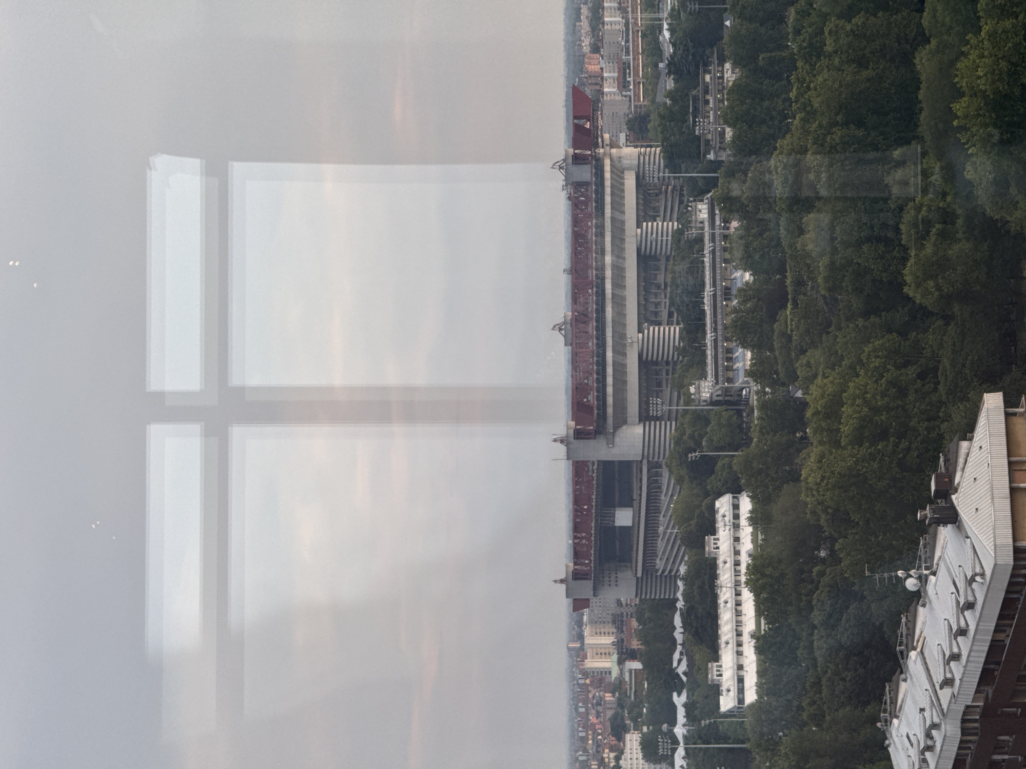 This is an outdoor, eye-level, medium shot photograph of a cityscape, taken from an elevated position, likely from a window. The image is characterized by reflections of windows on the left and right sides of the frame, which obscure portions of the scene.
Dominating the midground is the Stadio Giuseppe Meazza, also known as San Siro, a prominent football stadium in Milan, Italy. The stadium is distinguished by its circular, tower-like structures and a distinctive red roof. In front of the stadium, a long, white, rectangular building with multiple windows is visible, likely associated with the stadium complex or training facilities. Beyond the stadium, a dense urban sprawl of buildings extends into the distance, with a hazy, overcast sky overhead.
The foreground features the roofs of several apartment buildings, characterized by their grey, corrugated metal surfaces and ventilation structures. A large expanse of lush green trees fills the space between the foreground buildings and the stadium. The overall atmosphere is muted, suggesting either early morning or late afternoon lighting. No people are visible in the image.