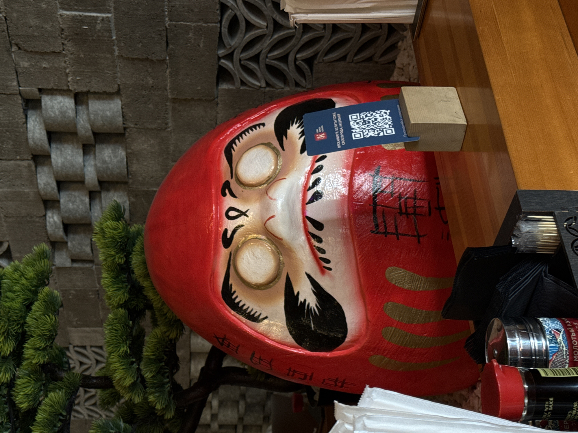 The image captures a close-up of a Daruma doll, a traditional Japanese doll, set against a background of gray textured blocks on the left and a wooden surface on the upper right. A small artificial bonsai tree is situated near the doll, adding a touch of greenery to the scene. A blue tag with a QR code and Russian text, "ОТСКАНИРУЙ, ЕСЛИ ТЫ ТОЖЕ, СВОЕГО РОДА, АКЦИОНЕР" (Scan if you're also, of sorts, a shareholder) is attached to the doll. To the right of the doll, there's a glimpse of condiments, including a container with the Russian words "МОЛОТЫЙ КУН" (Ground Kuy) visible on the label, and a bottle of soy sauce. This image was taken in the city of Krasnoyarsk, Russia.