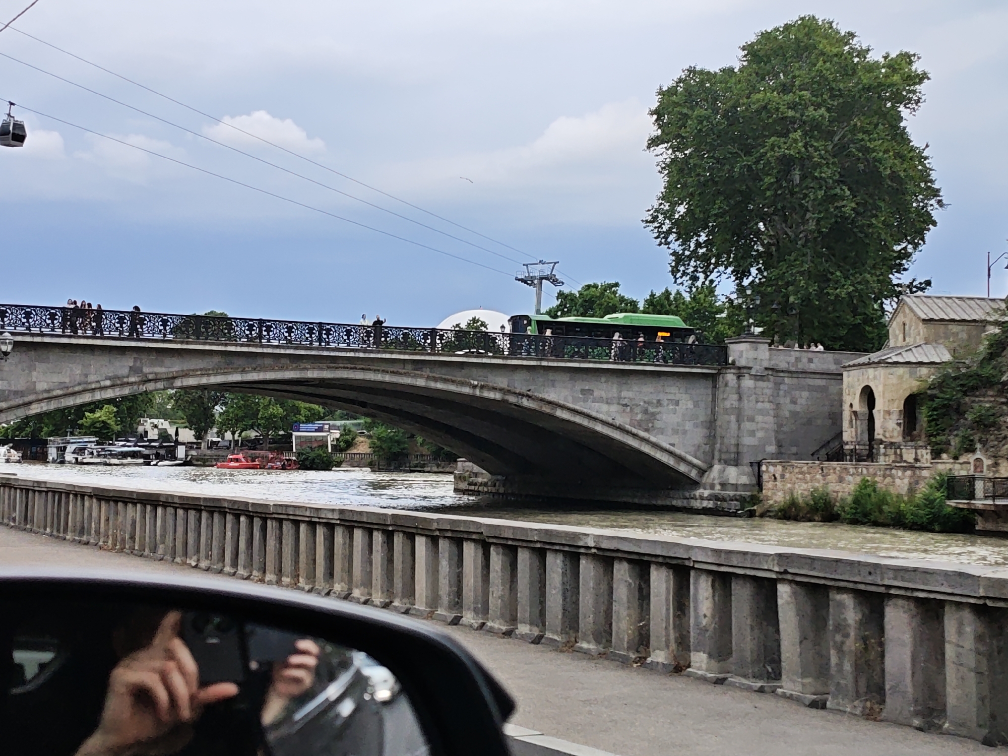 This media file captures a scenic view of a bridge spanning a river in Tbilisi, Georgia. The bridge, constructed of stone, features a wide arch and is lined with decorative railings. Several people are visible walking across the bridge, some pausing to look out. A green bus travels across the bridge, and further down the road, a cable car is seen suspended in the air against a cloudy sky.
The river below the bridge is wide and appears to be flowing moderately. Along the riverbank, a stone embankment with vertical pillars runs parallel to the water. Several boats are docked or moving on the river, including a bright red one. On the far bank, lush green trees, including a large, mature tree, frame the scene, and a portion of a building with an arched entryway is visible.
In the foreground, a reflection in a car's side mirror shows a person's hand and a smartphone, suggesting the image was taken from inside a vehicle. The overall impression is of a calm, overcast day, likely in the late morning or early afternoon, with the bustling activity of people and vehicles taking place around the picturesque urban landscape. There is no visible text in the image.