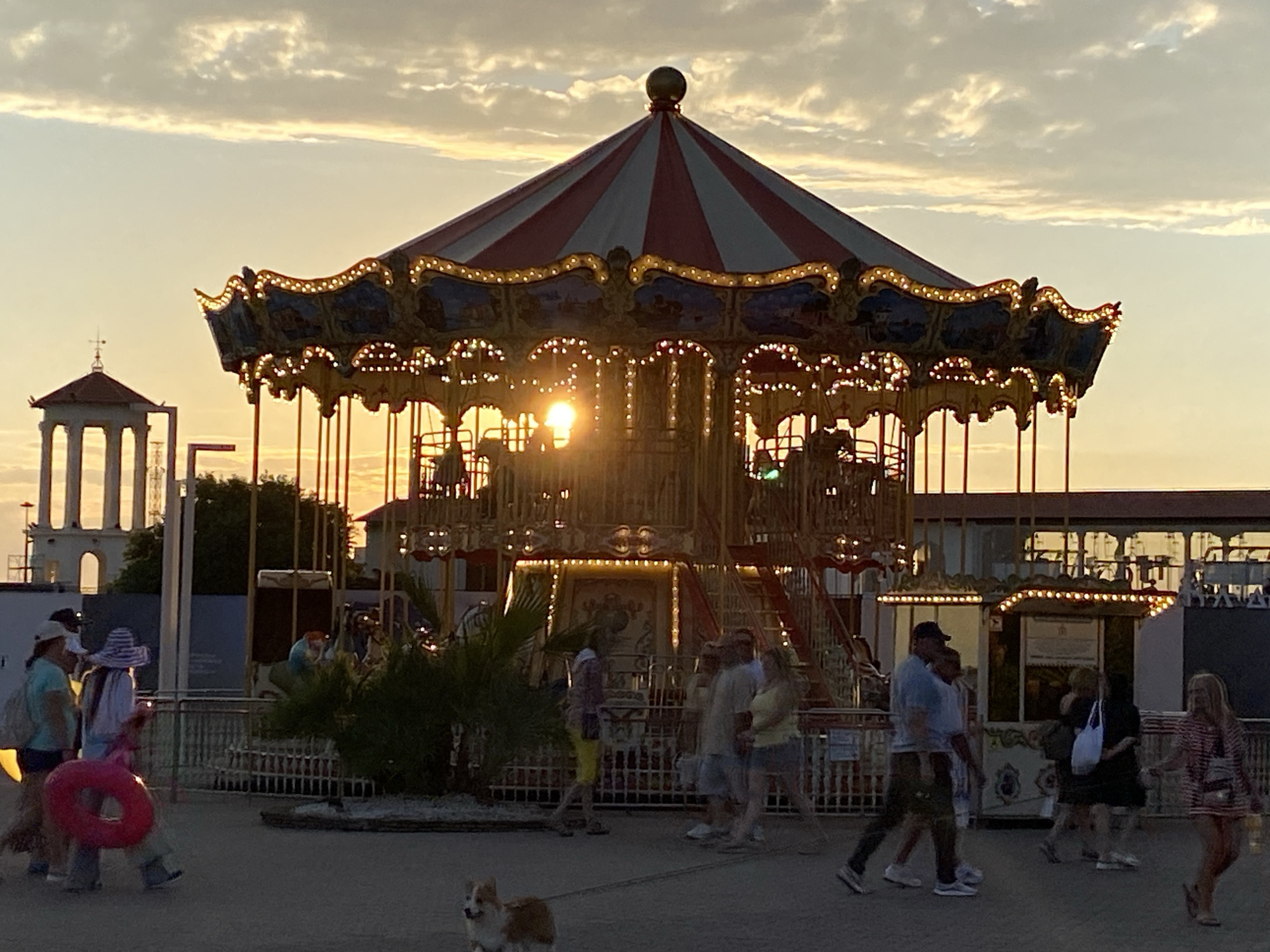 The image depicts a carousel at sunset in Sochi, Russia. The carousel, adorned with red and white stripes on its roof and illuminated by numerous lights, is the central focus. Several people are walking or standing near the carousel. In the foreground, a corgi is visible on a leash. In the background, a classical-style gazebo stands to the left, and various buildings line the perimeter of the amusement area. The sky is filled with clouds catching the golden light of the setting sun, creating a warm and atmospheric scene.