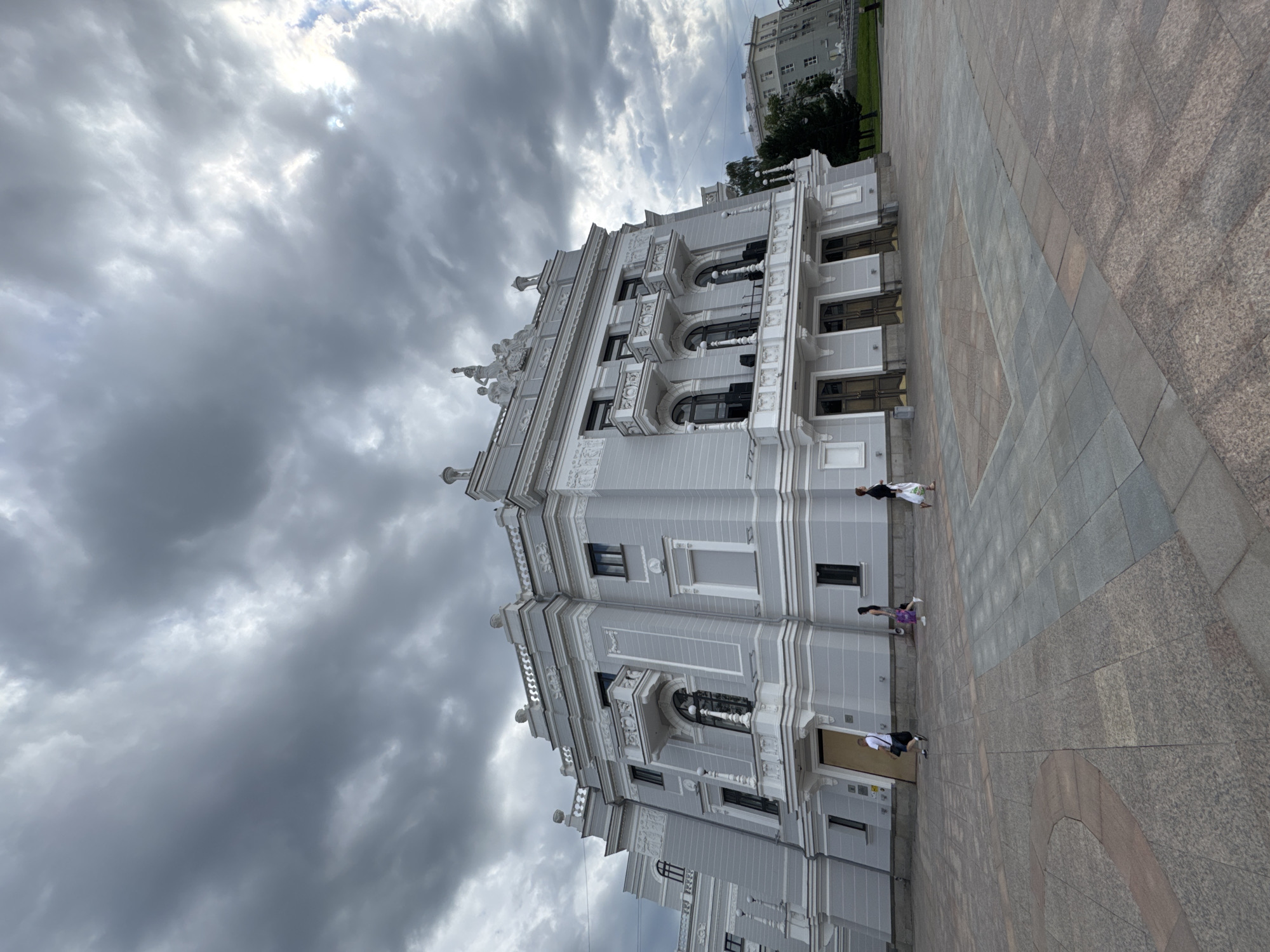 The image depicts a wide-angle view of the Sverdlovsk State Academic Theater of Musical Comedy in Yekaterinburg, Russia. The building, a grand edifice with a light gray facade, is centrally located in the frame. It features ornate architectural details, including multiple balconies with decorative railings, arched windows, and sculpted figures adorning the roofline. The building's facade is symmetrical, with imposing double doors leading to its entrance.
In the foreground, a large paved plaza is visible, laid with geometric patterns of gray and reddish-brown stone tiles. Three individuals are walking across the plaza towards the theater. To the left, a man in a white shirt and dark shorts walks with a black bag over his shoulder. In the middle, a woman in a colorful patterned dress walks with her back to the camera. To the right, another woman in dark clothing walks with a white bag in her hand. The sky above is heavily clouded, with dramatic, dark gray formations interspersed with patches of lighter gray and hints of diffused sunlight, suggesting an overcast or potentially stormy day. In the background, to the right of the main theater building, another, less ornate, multi-story building is visible, partially obscured by trees. The overall scene conveys a sense of urban civic space on a cloudy day.