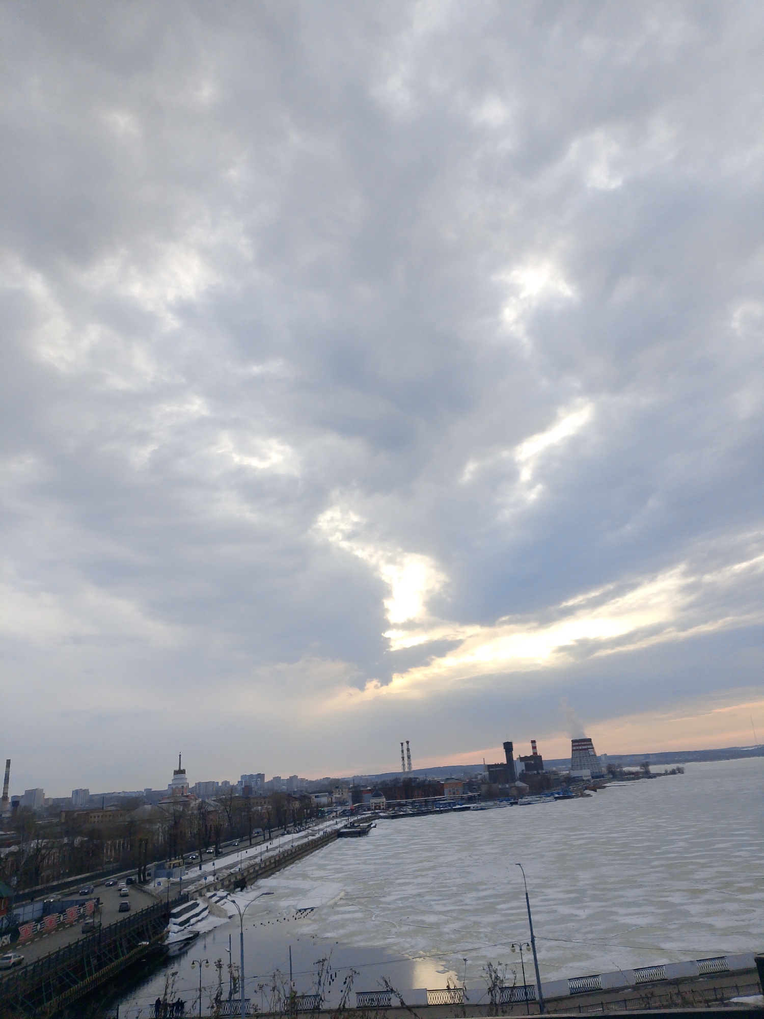 Here's a detailed description of the image:
**Environment:** The image depicts a cityscape under an overcast sky with scattered sunlight breaking through the clouds. The setting appears to be a waterfront area, likely a river or large lake, with patches of ice visible on the water's surface.
**Location:** The location is the city of Izhevsk, Russia. This is based on the recognizable cityscape elements such as the dam, Central Square, Udmurt State University, and the chimneys of Izhevsk TPP-1 and TPP-2.
**Subjects & Objects:**
* **Waterfront:** A paved walkway or embankment runs along the edge of the water.
* **Buildings:** A mix of low-rise and high-rise buildings form the urban skyline.
* **Industrial Structures:** Chimneys or smokestacks suggest the presence of an industrial area, power plant or thermal station near the water.
* **Vehicles:** Cars and possibly buses or trucks are visible on the roads along the embankment.
* **People:** Some figures are standing or walking near the walkway.
* **Lighting Fixtures:** Streetlights are present along the waterfront promenade.
**Actions & Interactions:**
* Vehicular traffic moving along the roads.
* Pedestrians strolling along the walkway.
* Industrial structures emitting smoke.
**Overall Impression:** The image conveys a sense of urban activity coexisting with a partially frozen body of water under a cloudy sky. The lighting indicates either dawn or dusk. The scene depicts a blend of urban, natural, and industrial elements.